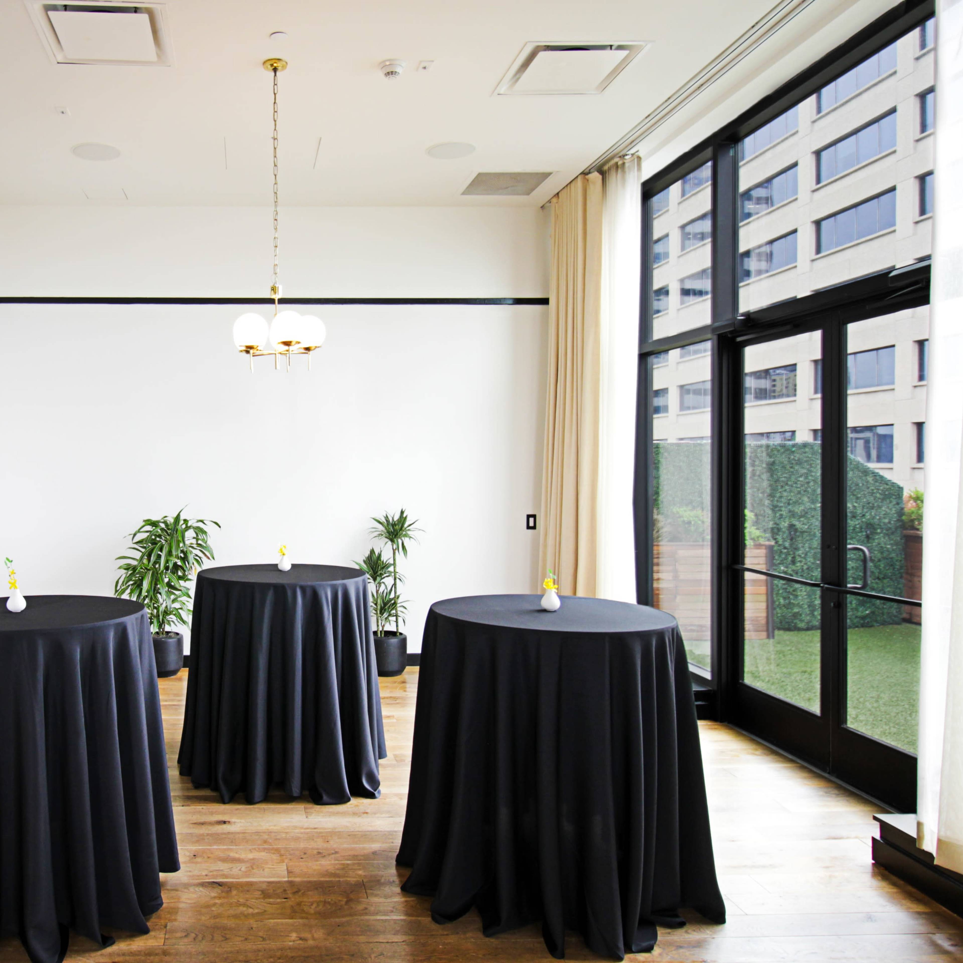 A bright room with three round tables covered in black tablecloths, arranged near large windows overlooking a green area.