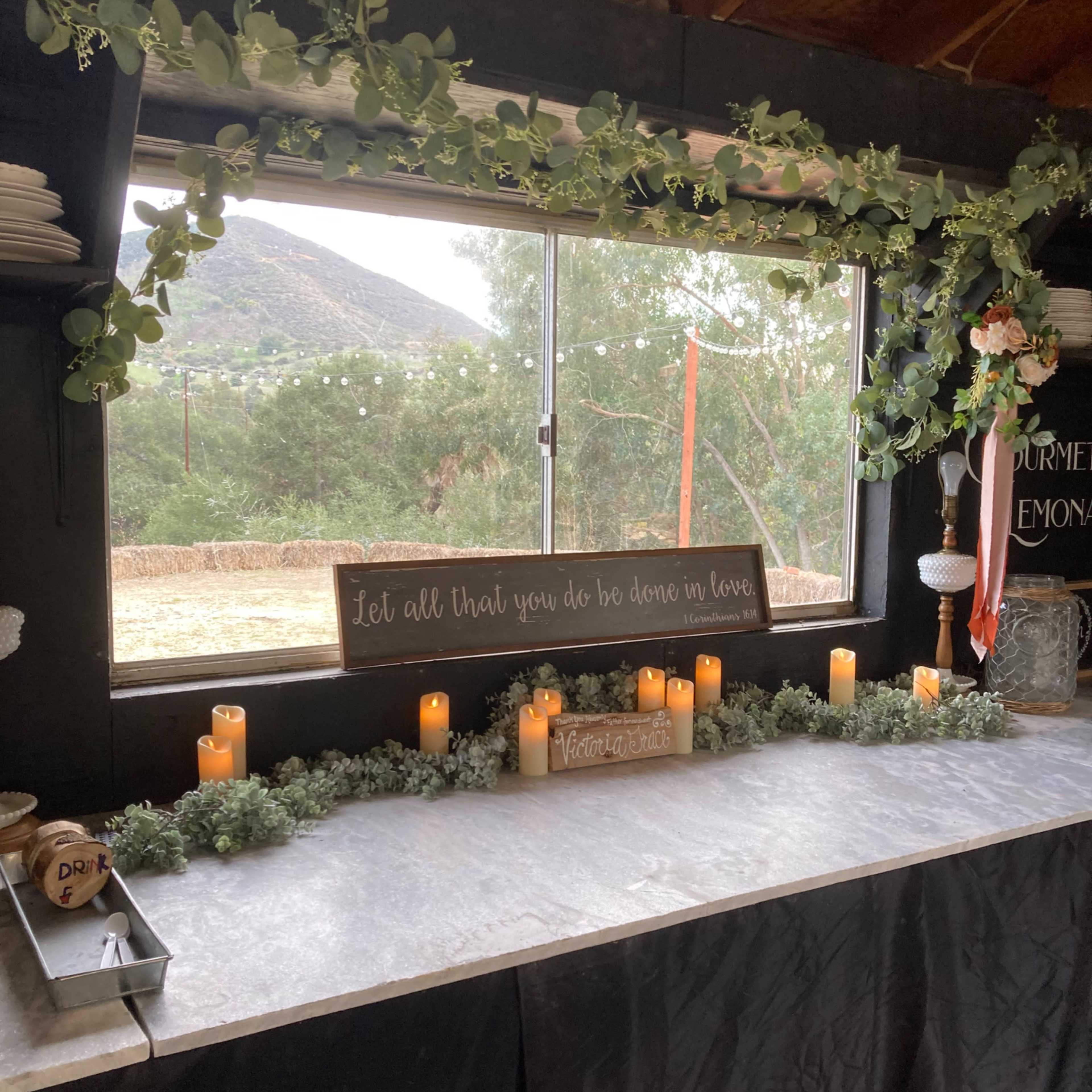 A decorated counter with candles, greenery, and a sign set against a backdrop of a window looking out at a hillside.