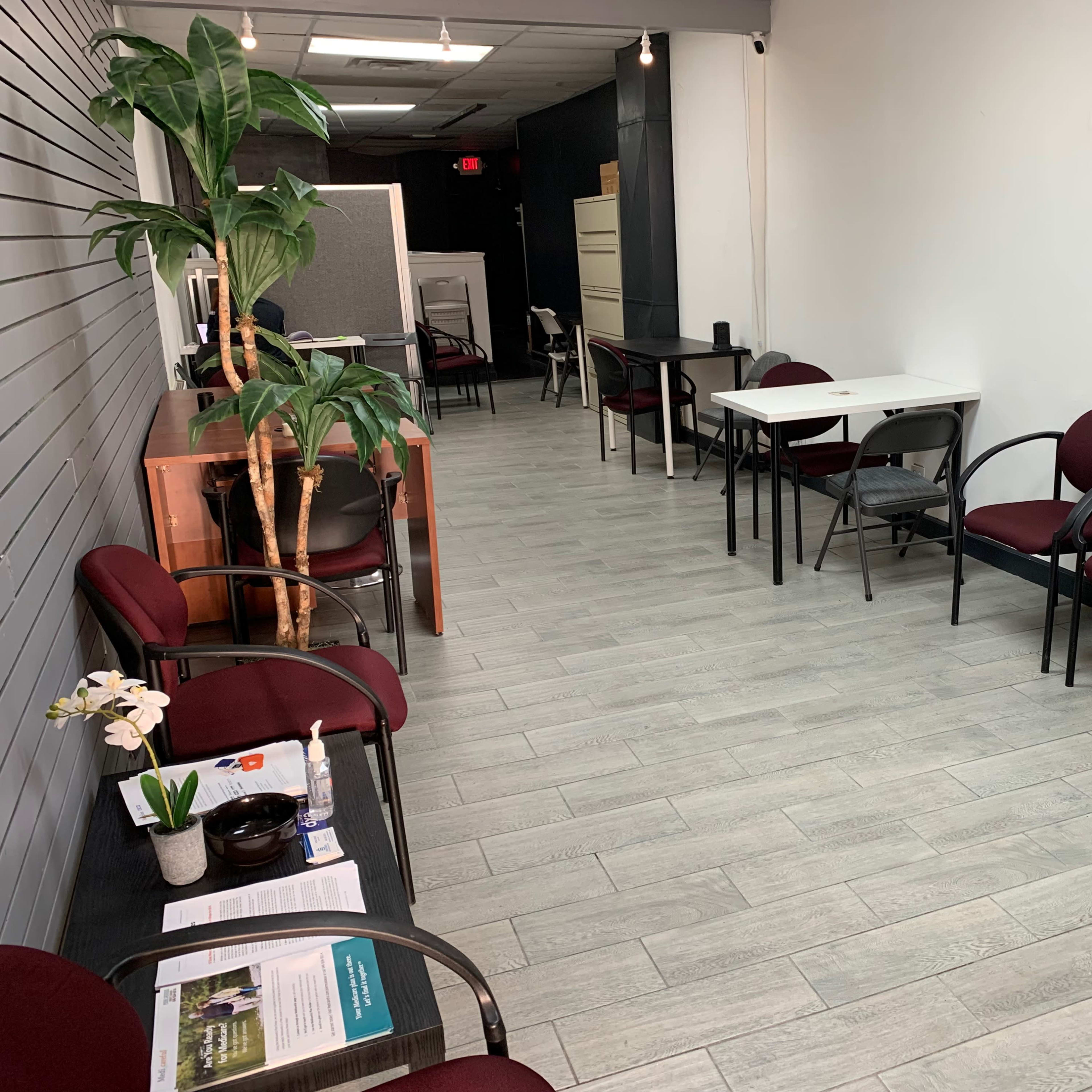 A waiting area with several maroon chairs, a potted plant, and a table, set against a light-colored wall and gray tile floor.