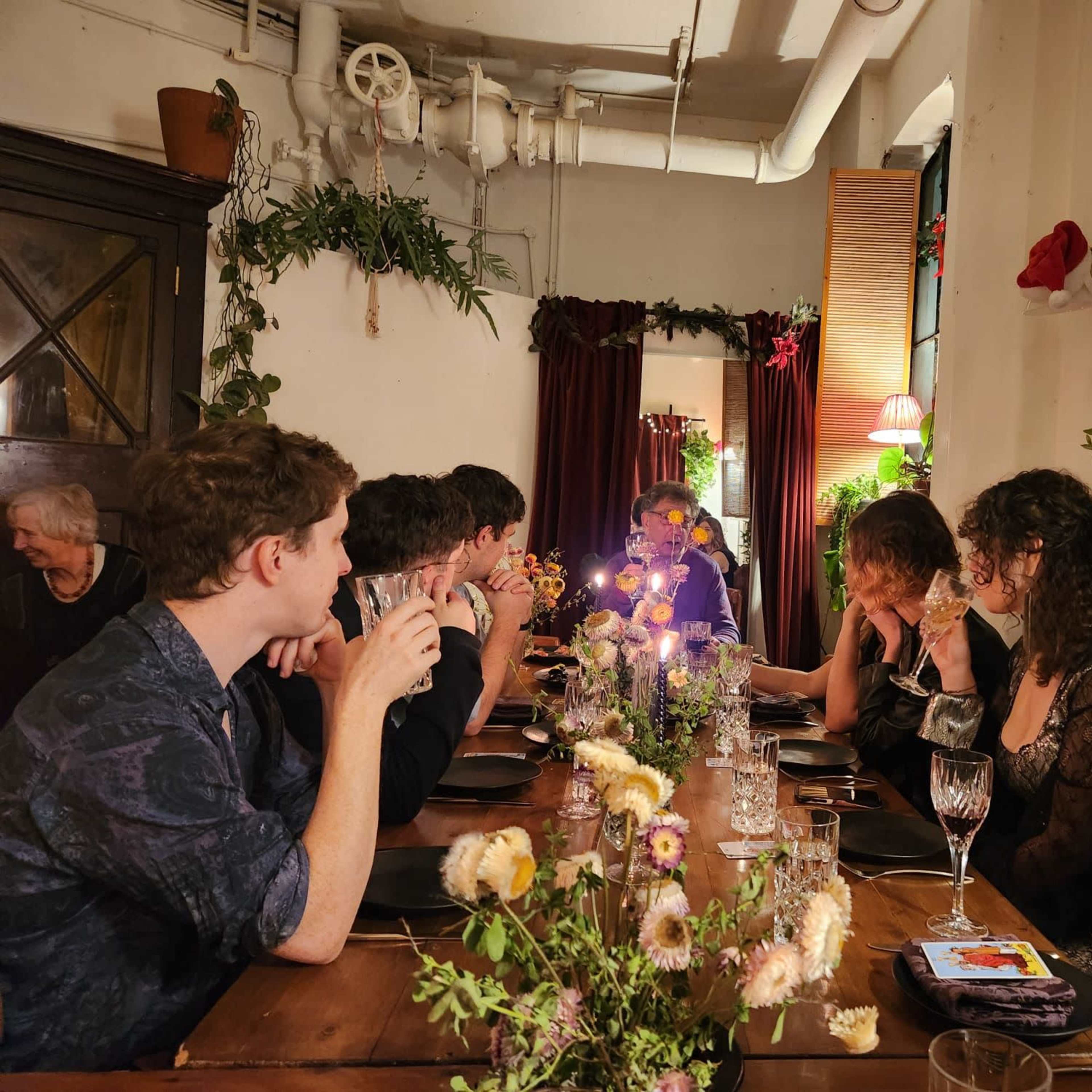 A group of people sits around a wooden dining table adorned with flowers and candles in a warmly lit room.