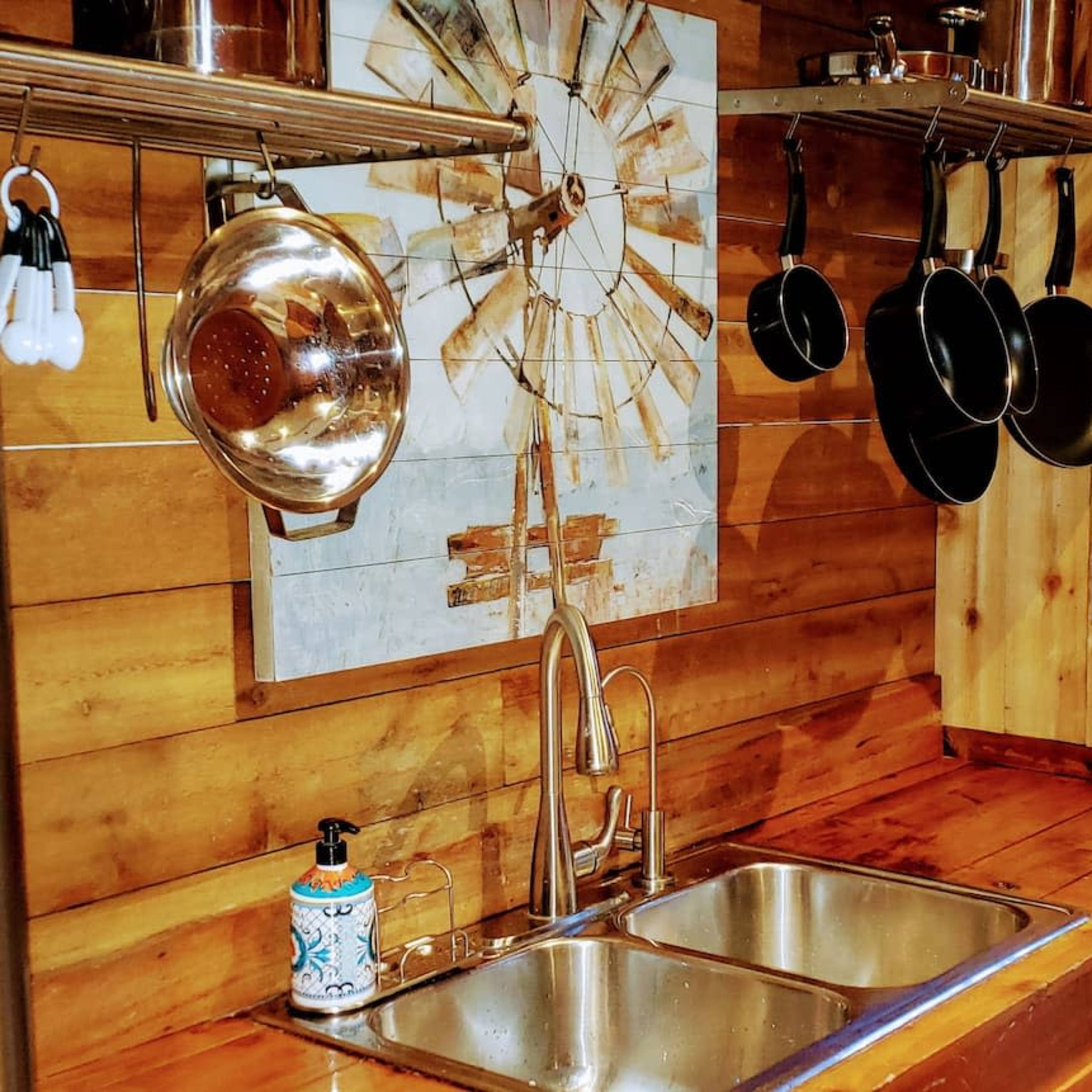A wooden kitchen with a double sink, hanging pots and pans, a decorative wall piece, and a glass door leading to an outdoor area with greenery.