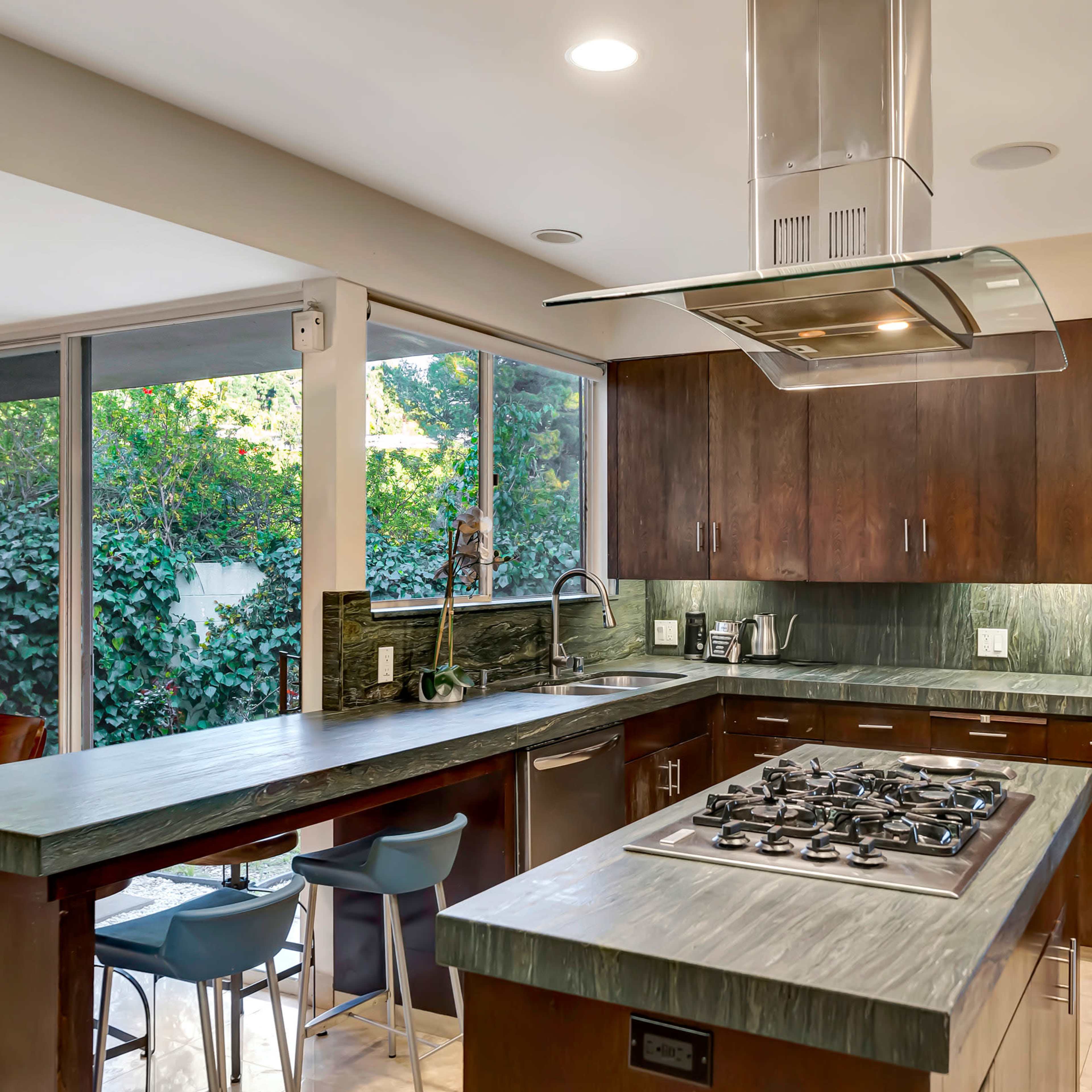 A modern kitchen with wooden cabinetry, a large island with a gas cooktop, and large windows that provide natural light and a view of greenery outside.