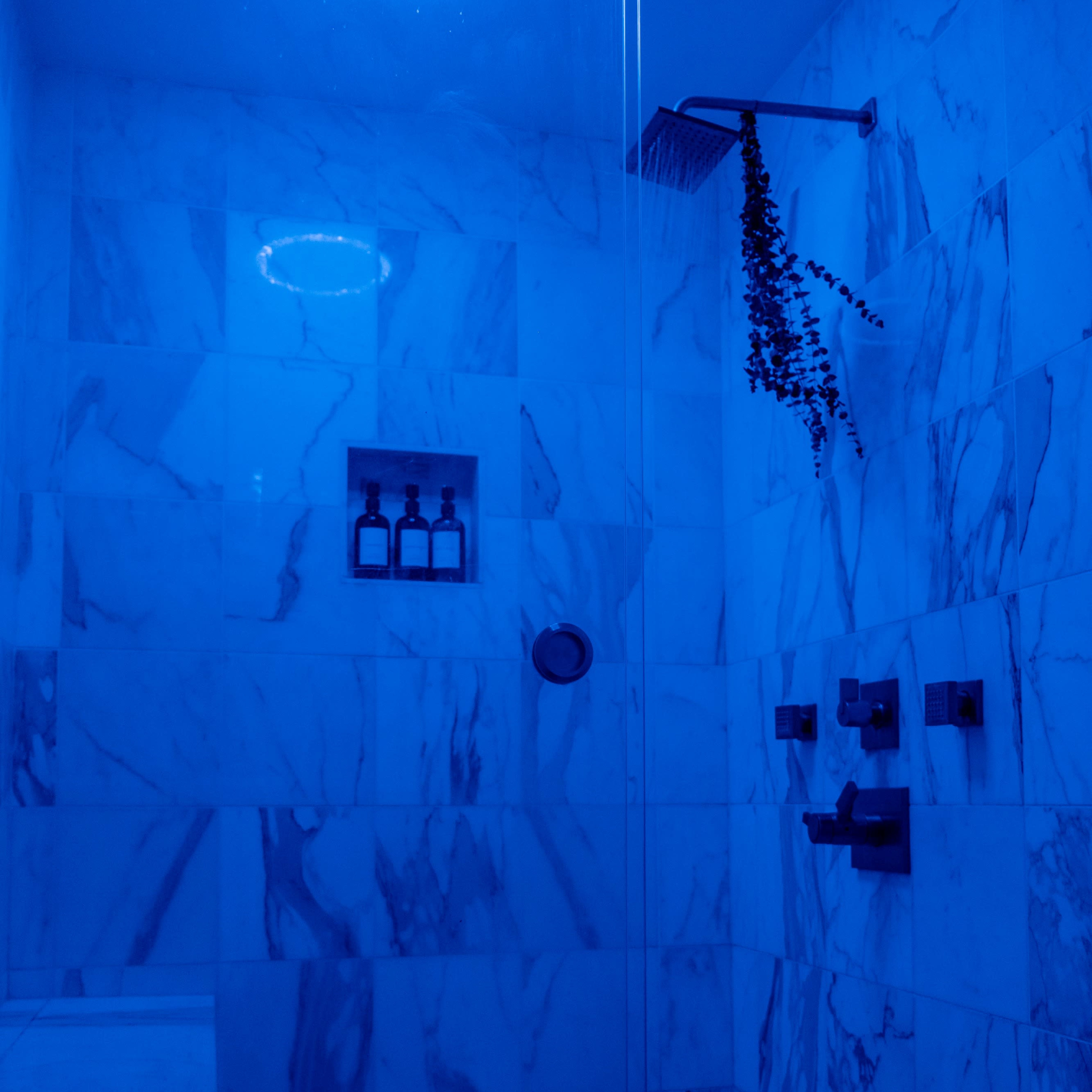 A modern shower enclosure features blue lighting, marble walls, and a wall-mounted showerhead with a shelf for toiletries.