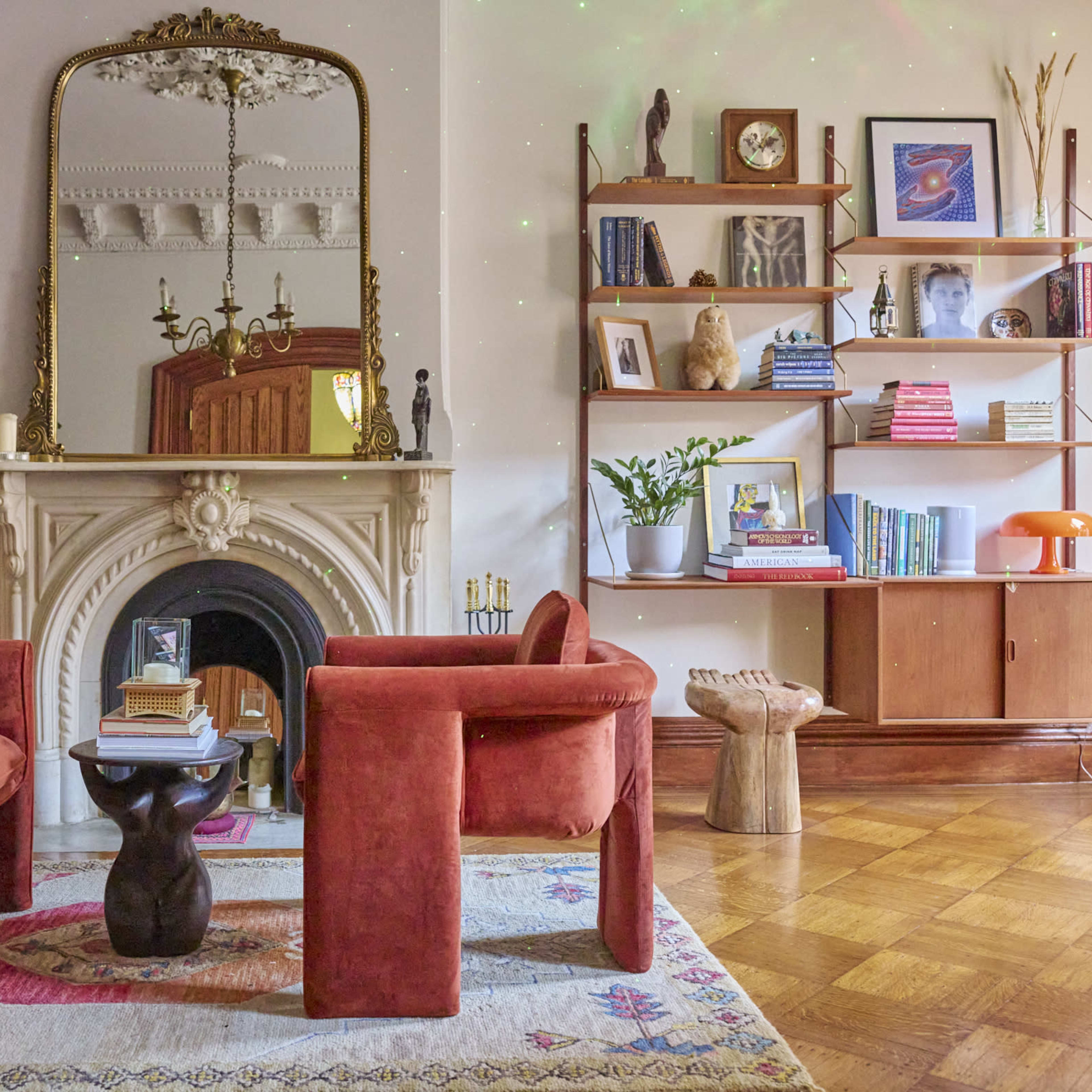 A cozy living room features two red armchairs facing a small black table, with a marble fireplace and a shelf filled with books and decorative items in the background.