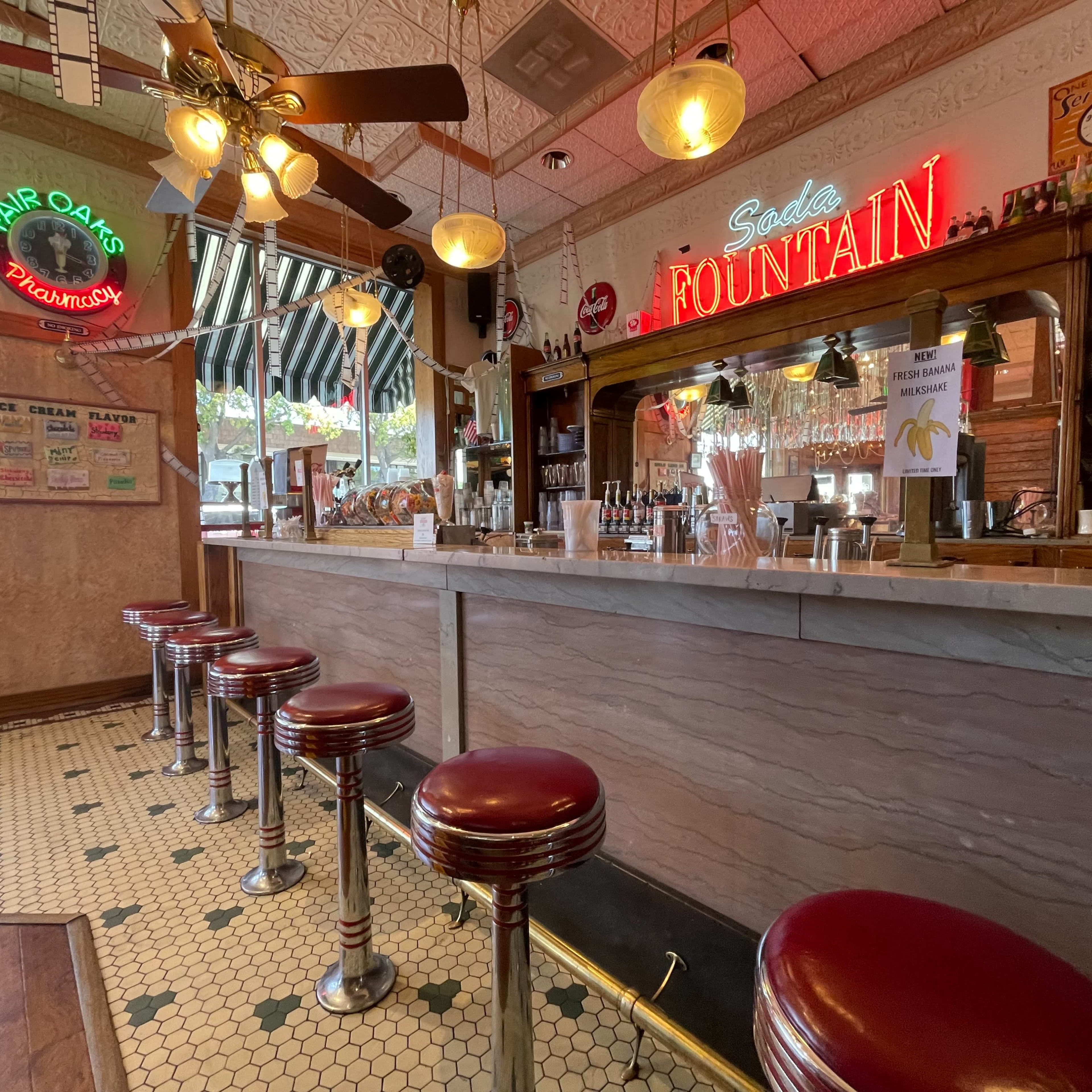 A vintage soda fountain with red barstools, a marble counter, and retro signage in a well-lit diner setting.