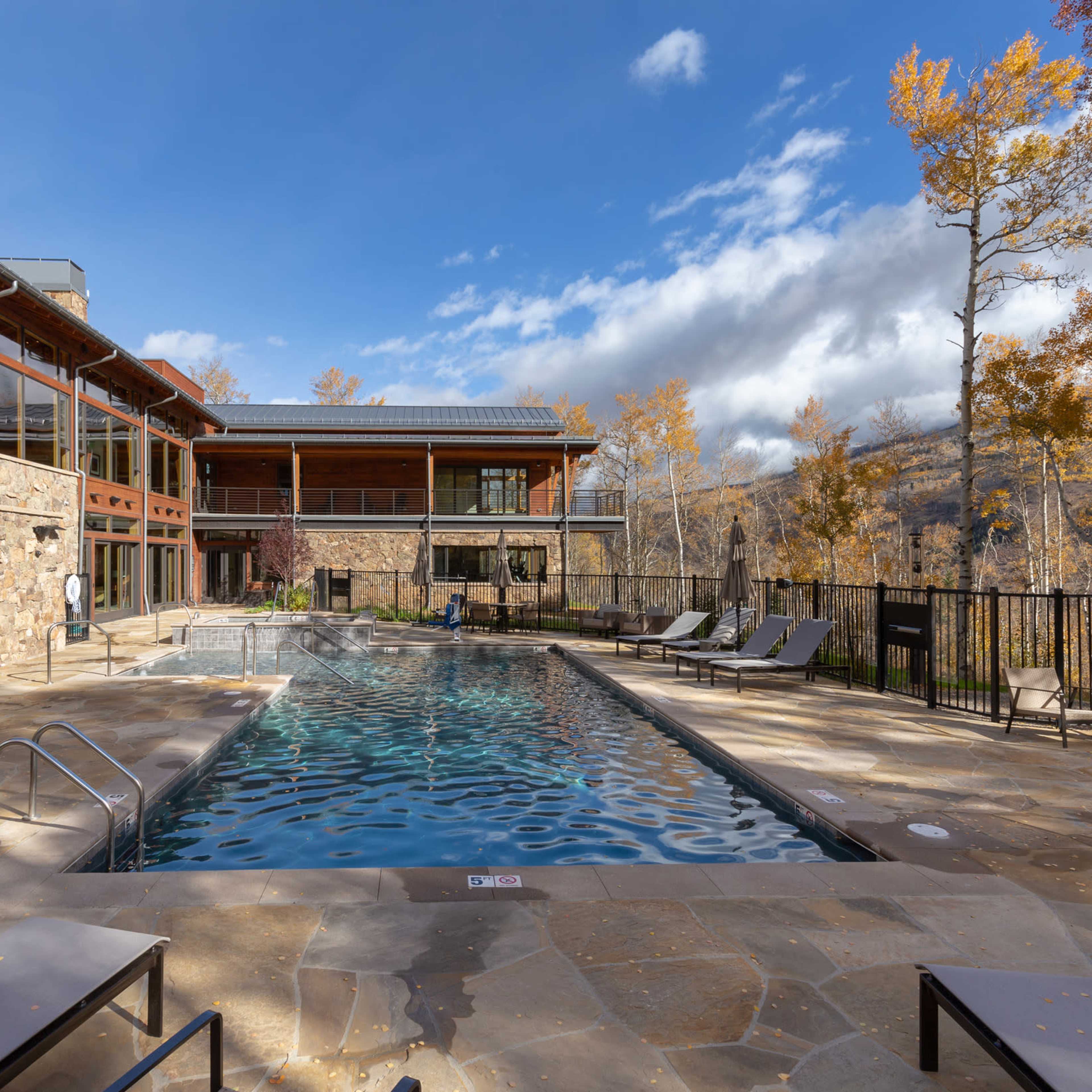 A spacious outdoor pool area surrounded by lounge chairs, with a modern building and autumn trees in the background.