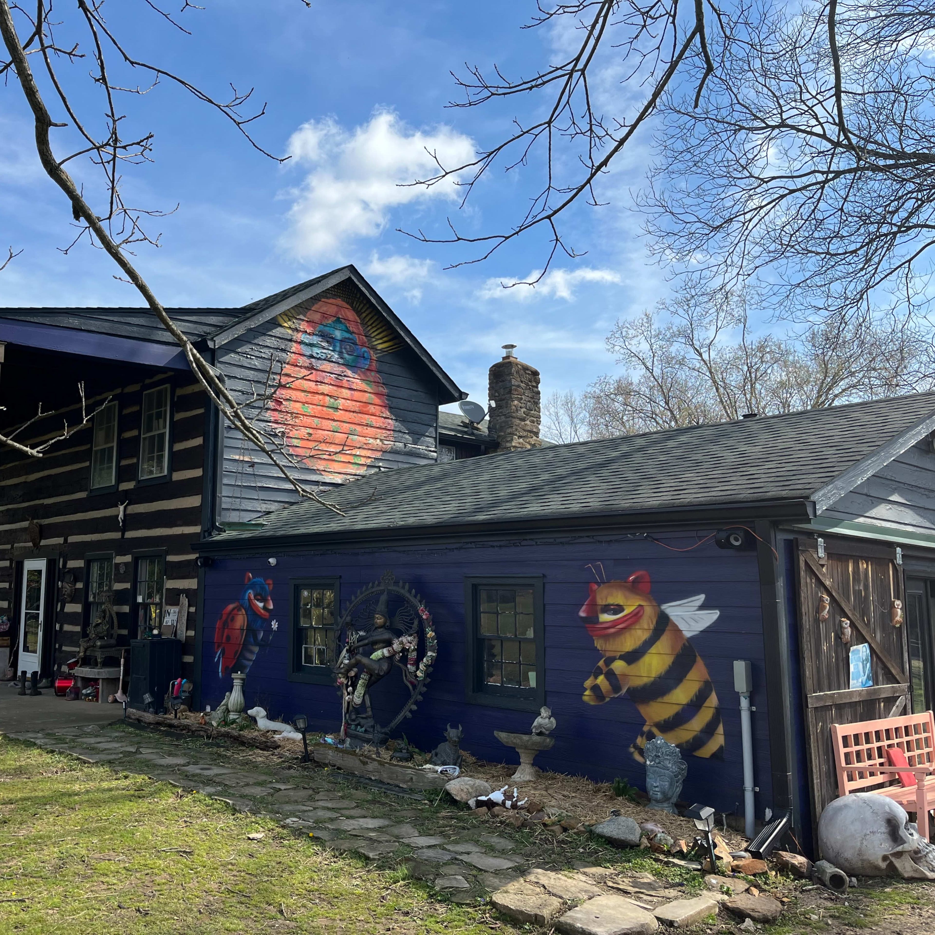 A wooden house features a colorful mural of a bee and a bird on its side, with a grassy yard in front and trees overhead.
