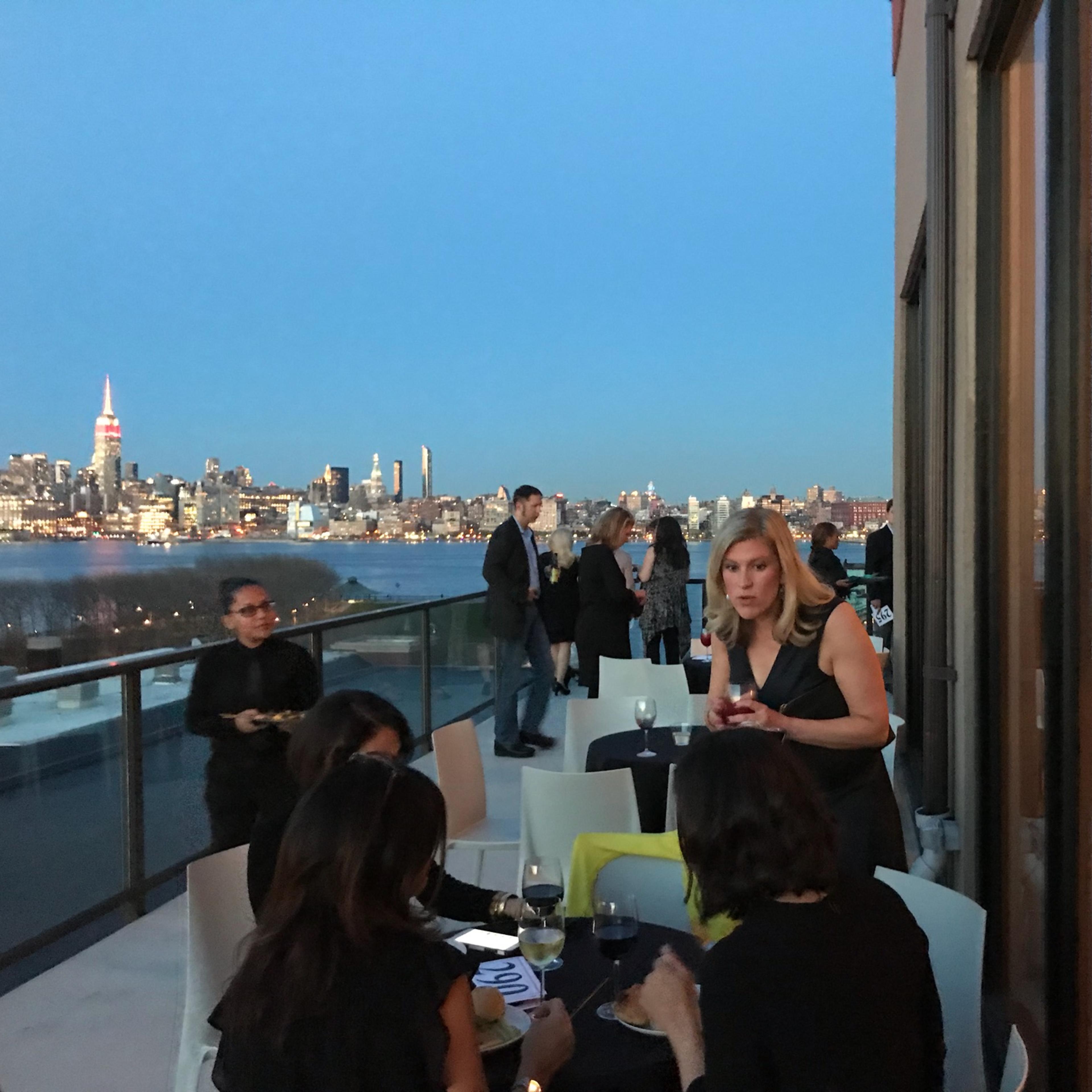 A group of people socializes on a rooftop terrace overlooking the Manhattan skyline at dusk.