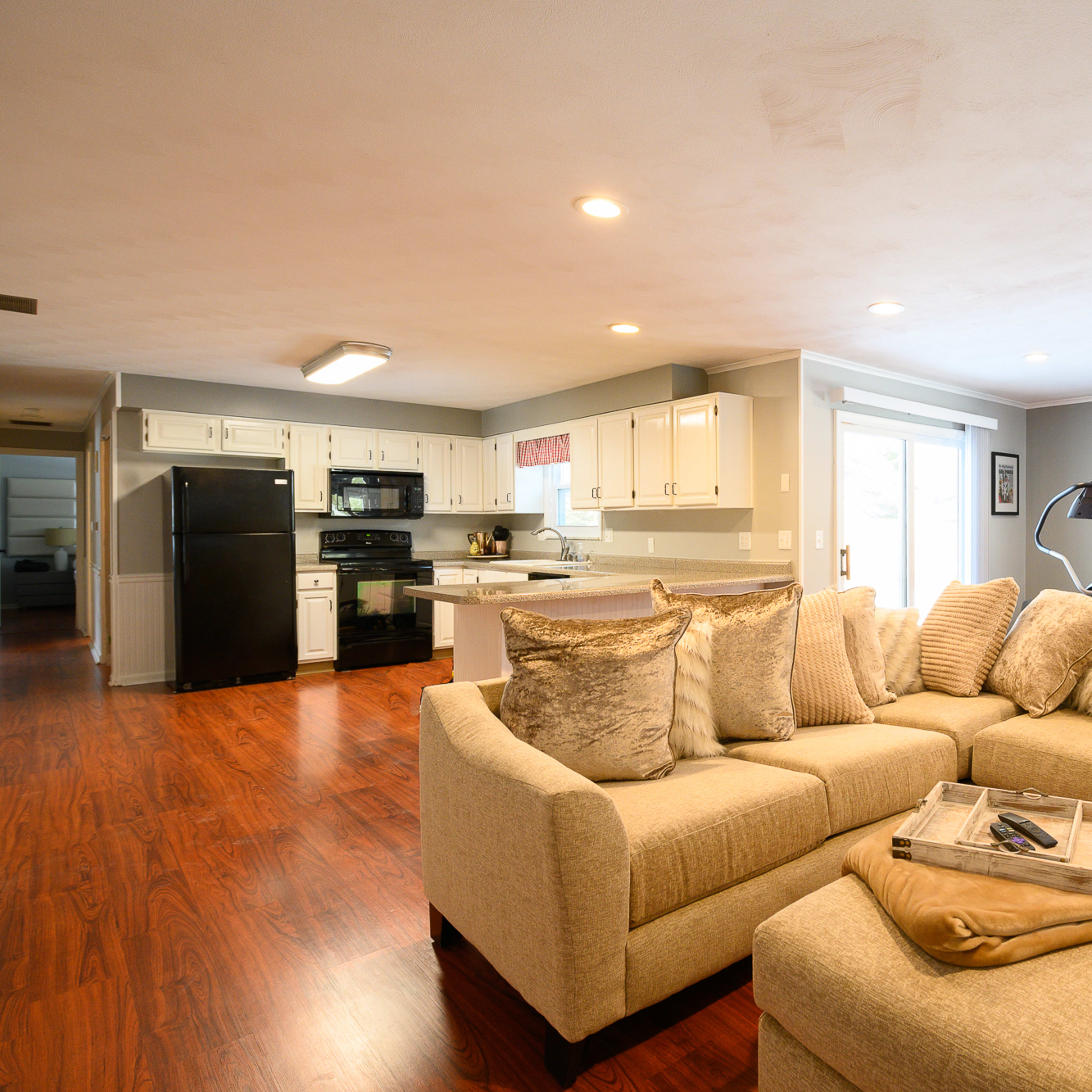 A light-colored sectional sofa with decorative pillows is arranged in a spacious living room that features a kitchen area and large windows.