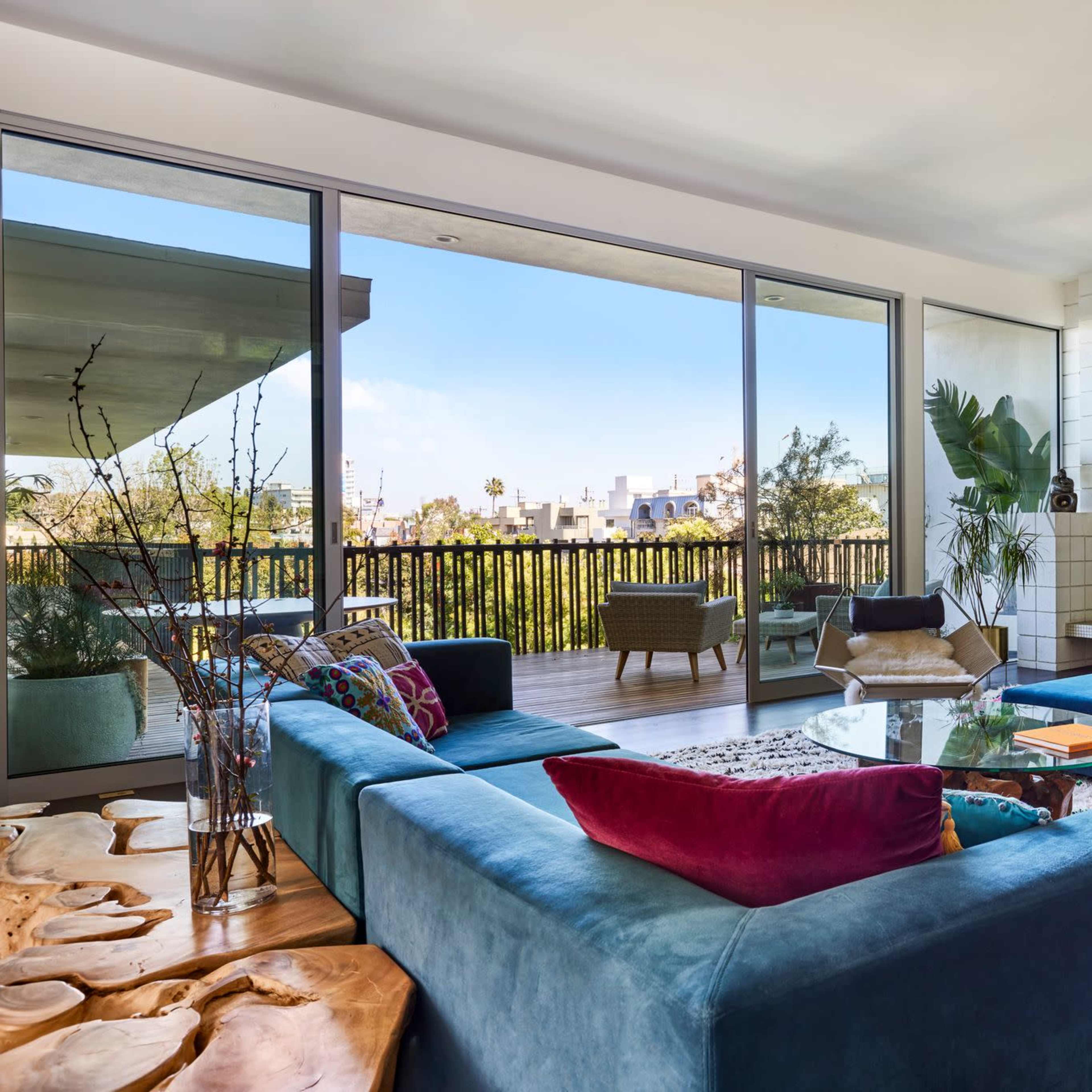 A modern living room featuring a blue sectional sofa, a coffee table, large windows providing a view of an outdoor balcony, and various decorative elements.