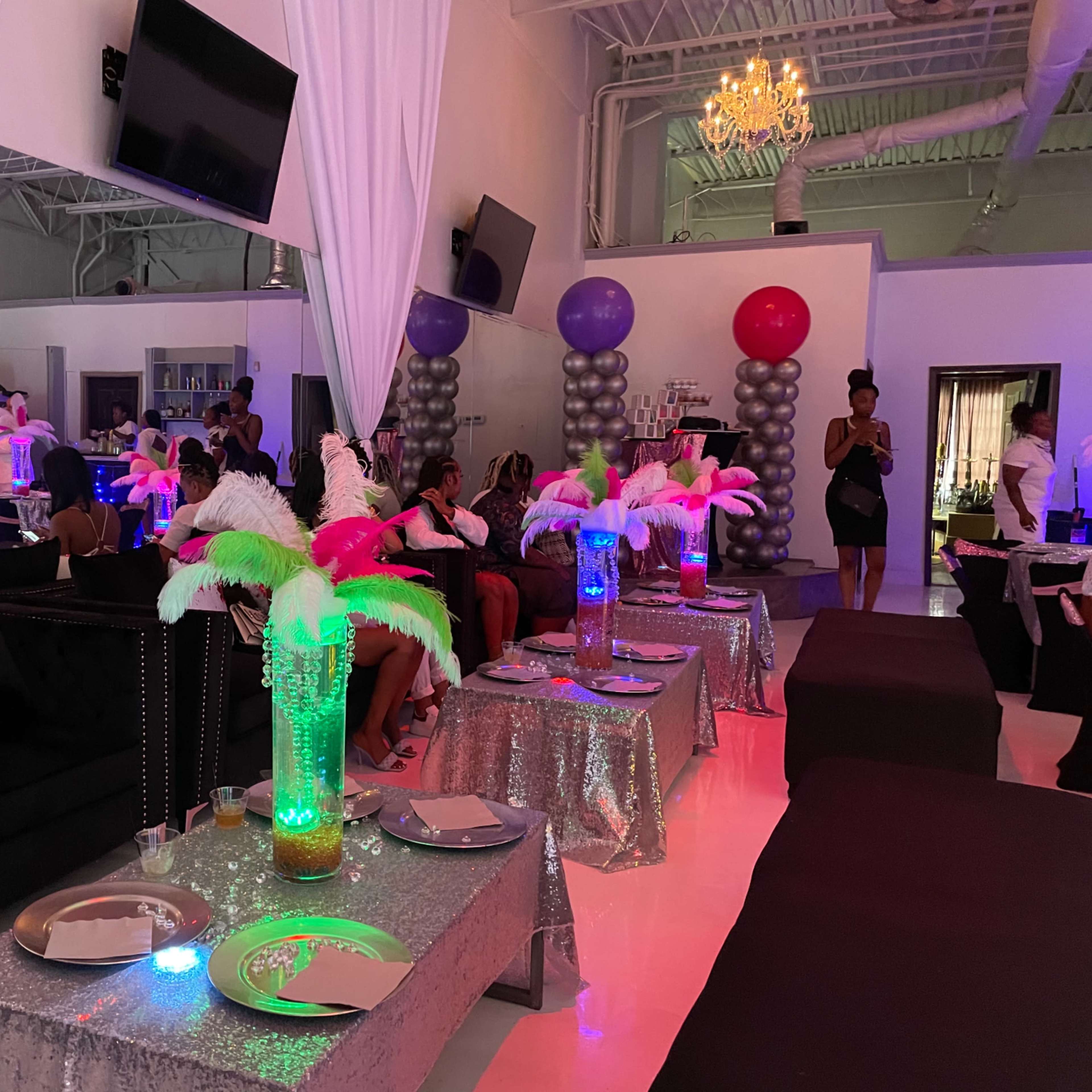 A brightly decorated event space with tables covered in sequined cloths, feather centerpieces, and guests seated around in a festive atmosphere.