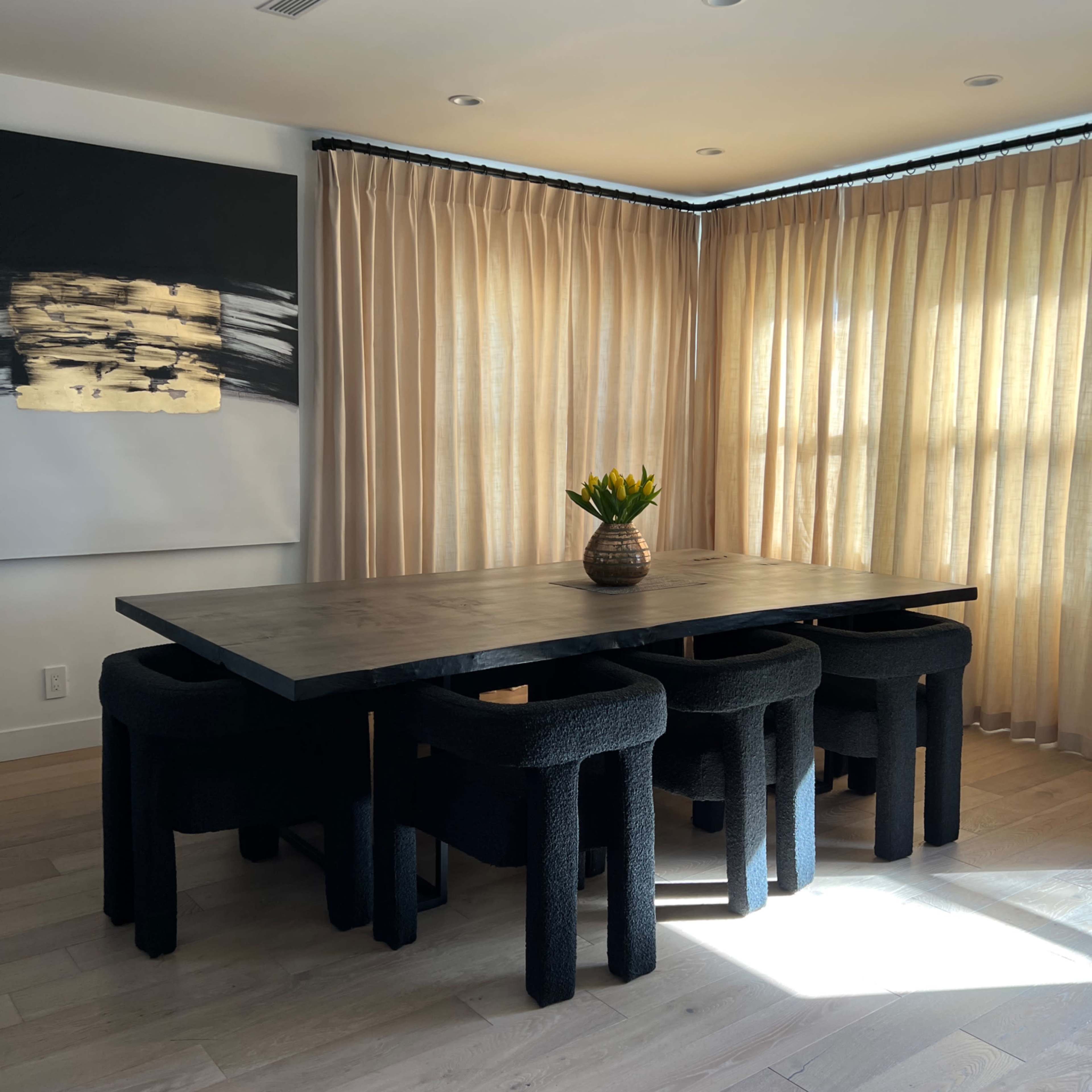A modern dining area features a large table surrounded by black upholstered stools, with a vase of flowers and sheer curtains filtering natural light.