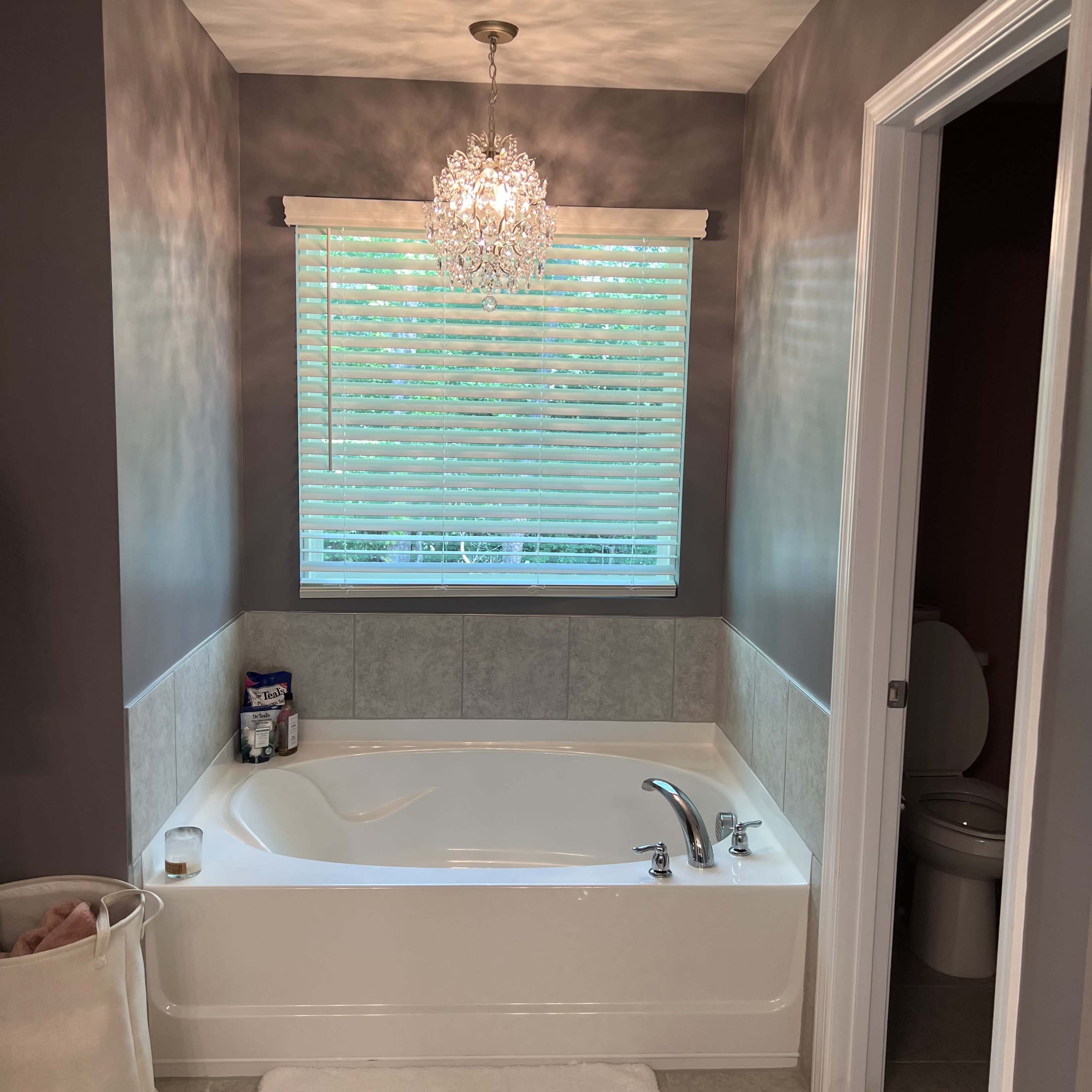 A bathroom with a white bathtub, a chandelier above it, and a window with blinds allowing natural light.
