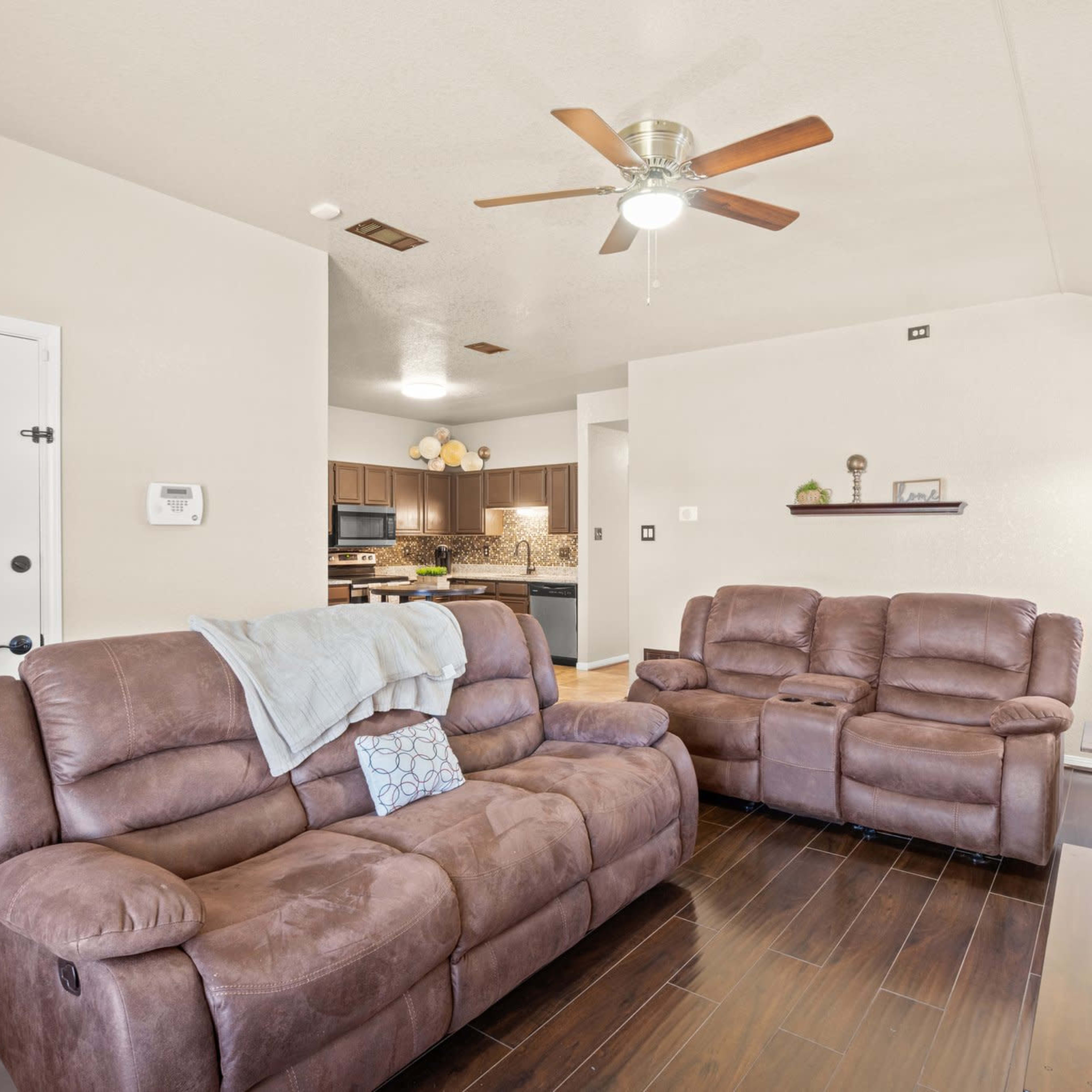 A living room with brown reclining sofas, a ceiling fan, and a view of a kitchen in the background.