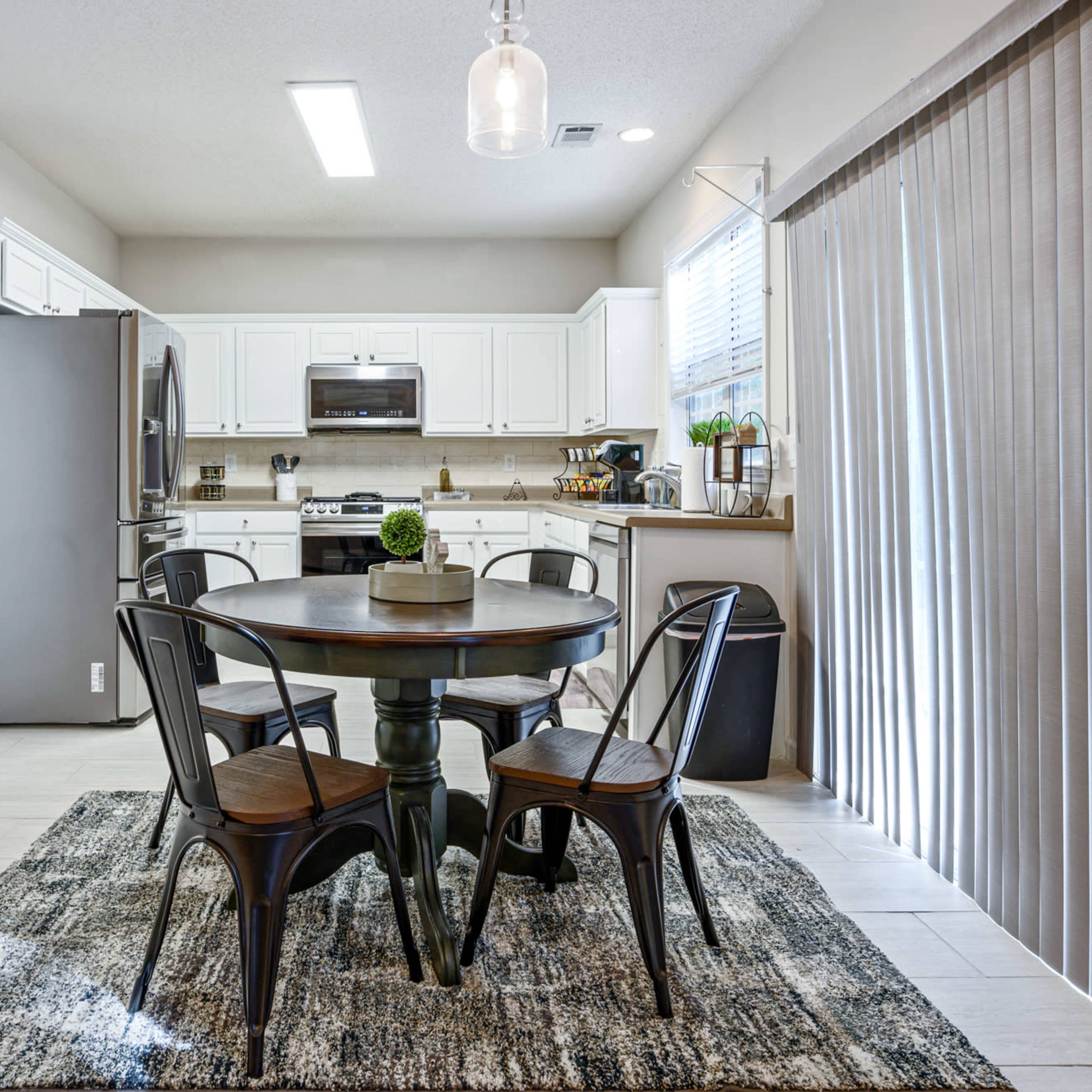 A modern kitchen features a round wooden table with four chairs, stainless steel appliances, and a sliding door with vertical blinds.