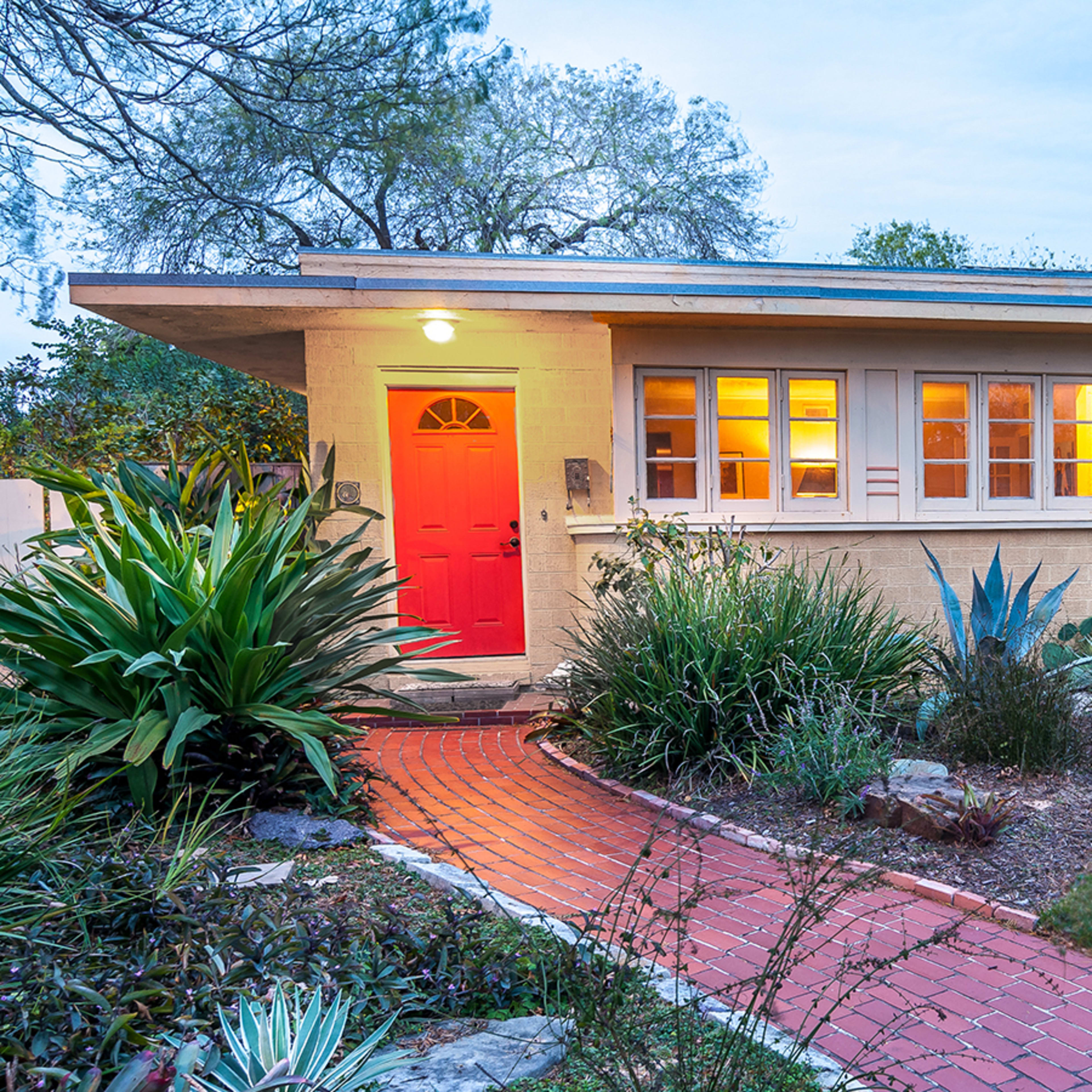 A modern house with a red door sits at the end of a brick pathway, surrounded by various shrubs and palm trees.