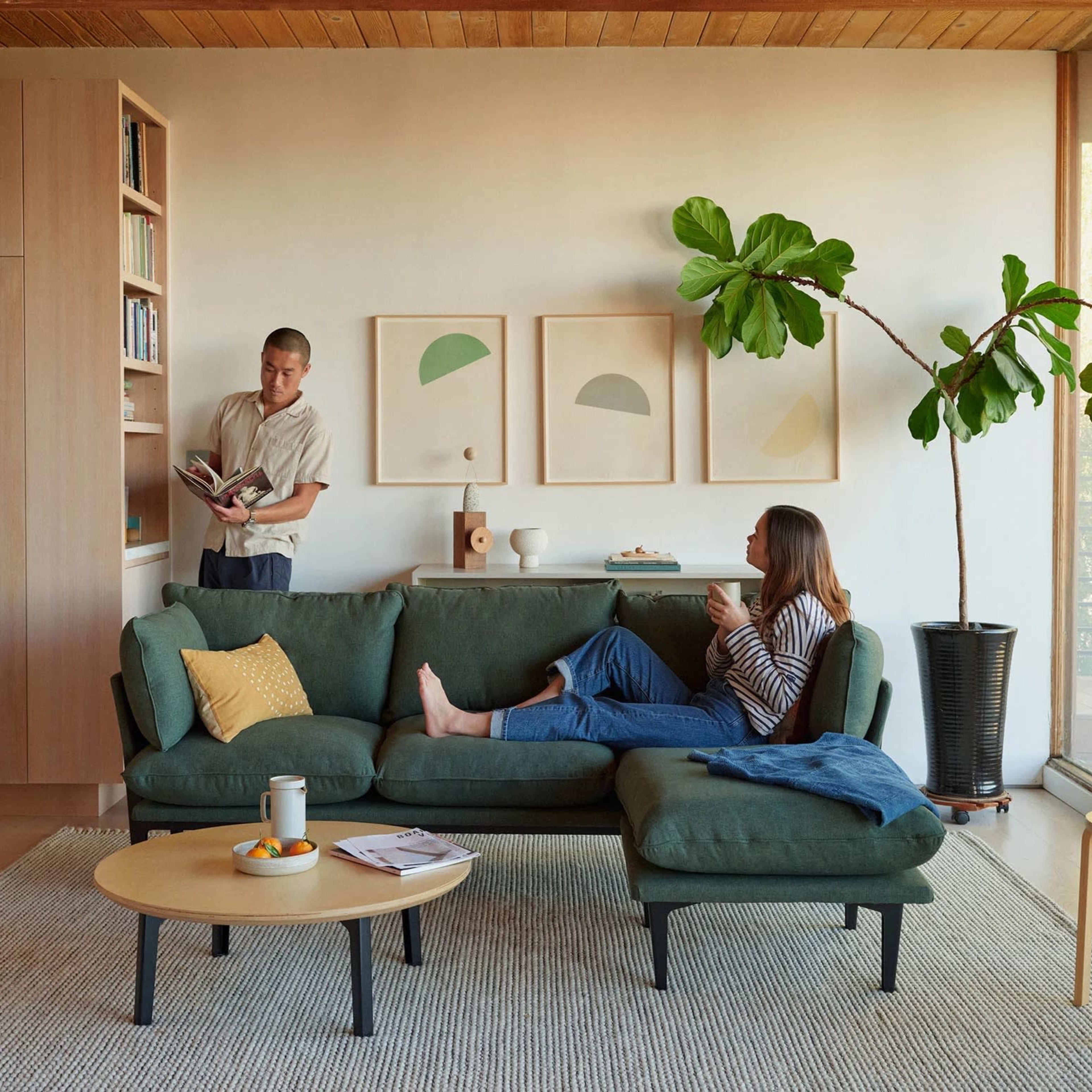 A man reads a book on a bookshelf while a woman sits on a green sofa, holding a drink.