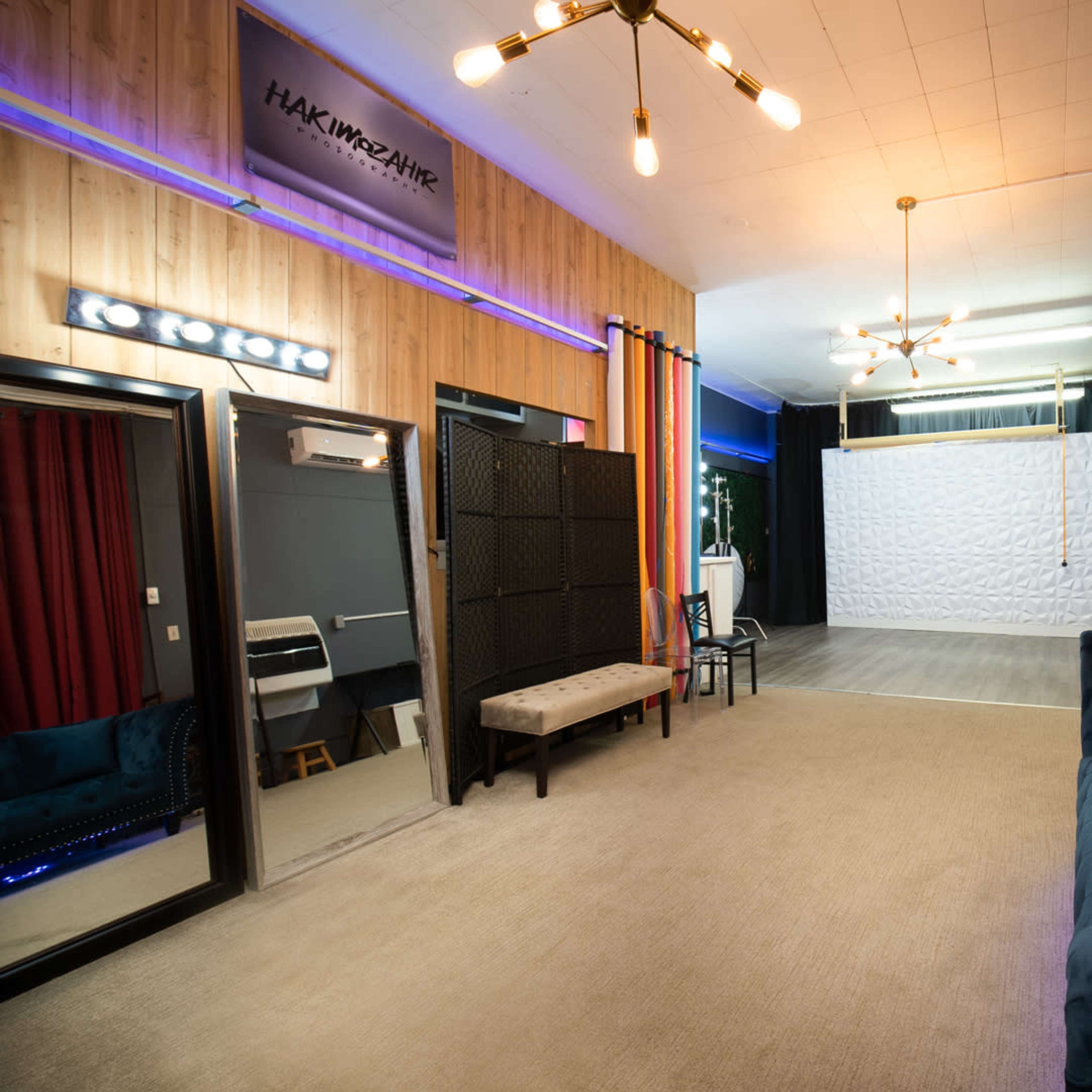 A spacious room with a blue couch, a mirror, styling chairs, and a white backdrop, illuminated by overhead lighting.