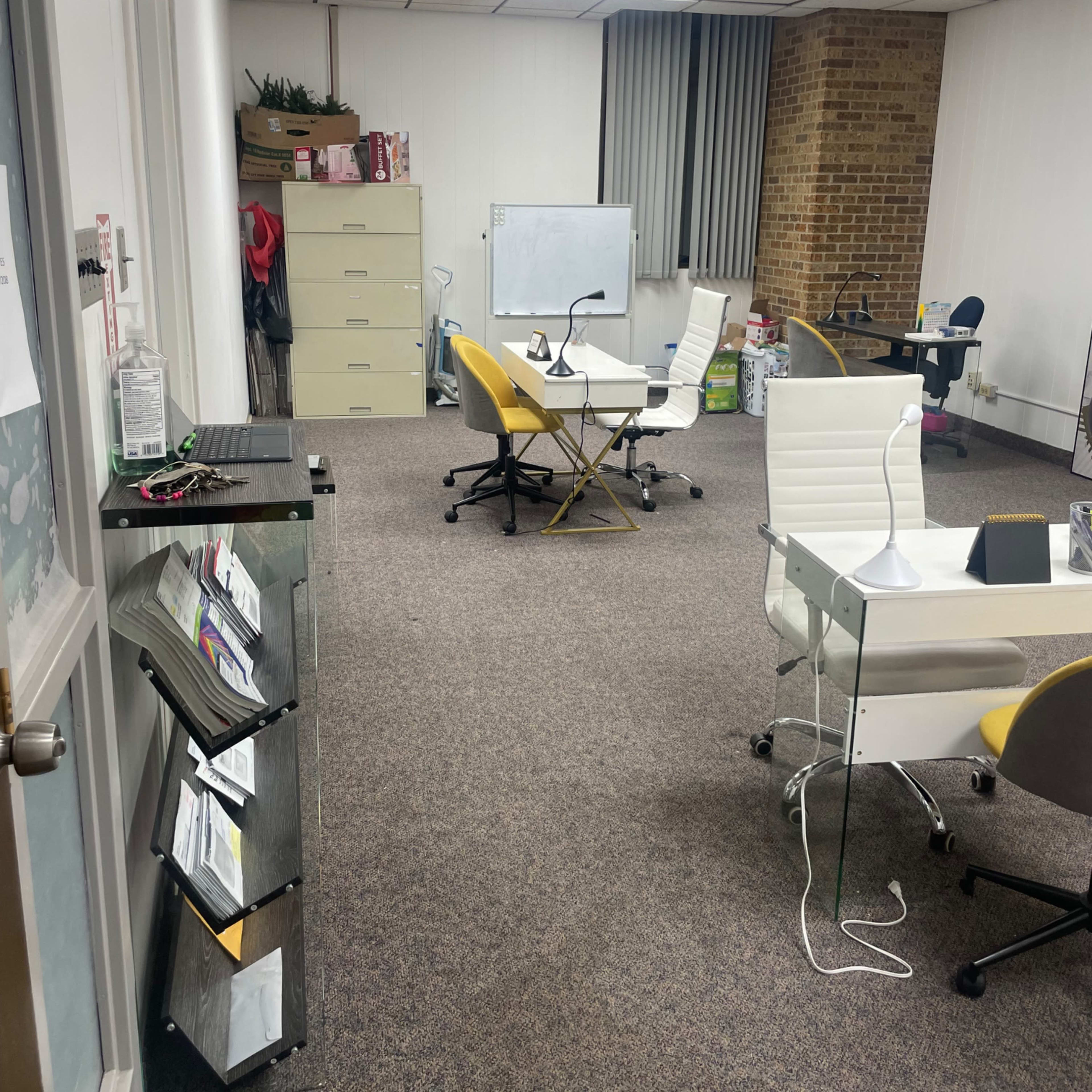 An office space with desks, chairs, a filing cabinet, and a whiteboard in a room featuring brick walls and carpeting.