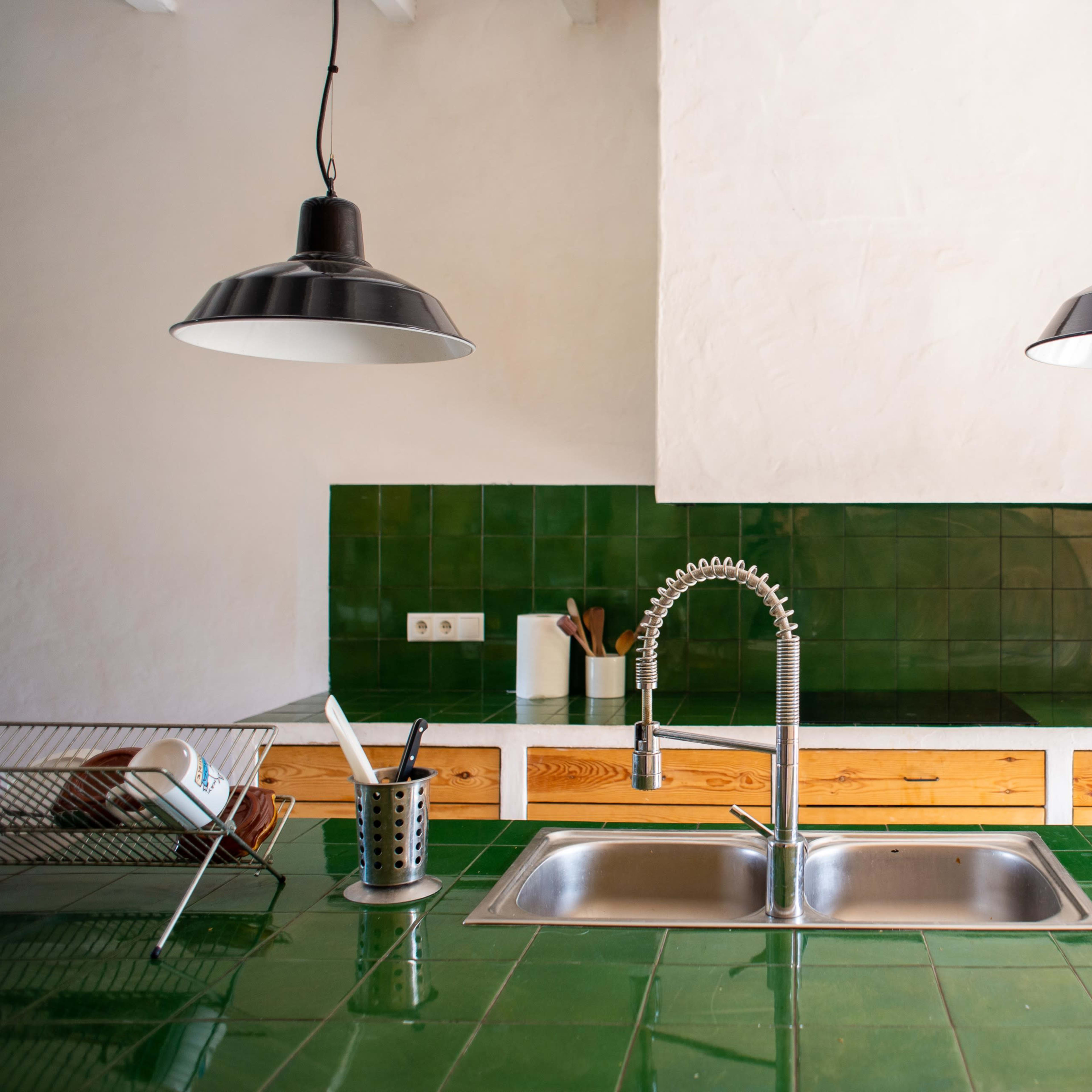 A modern kitchen features a green tiled countertop with a stainless steel sink, wooden cabinets, and two black pendant lights.