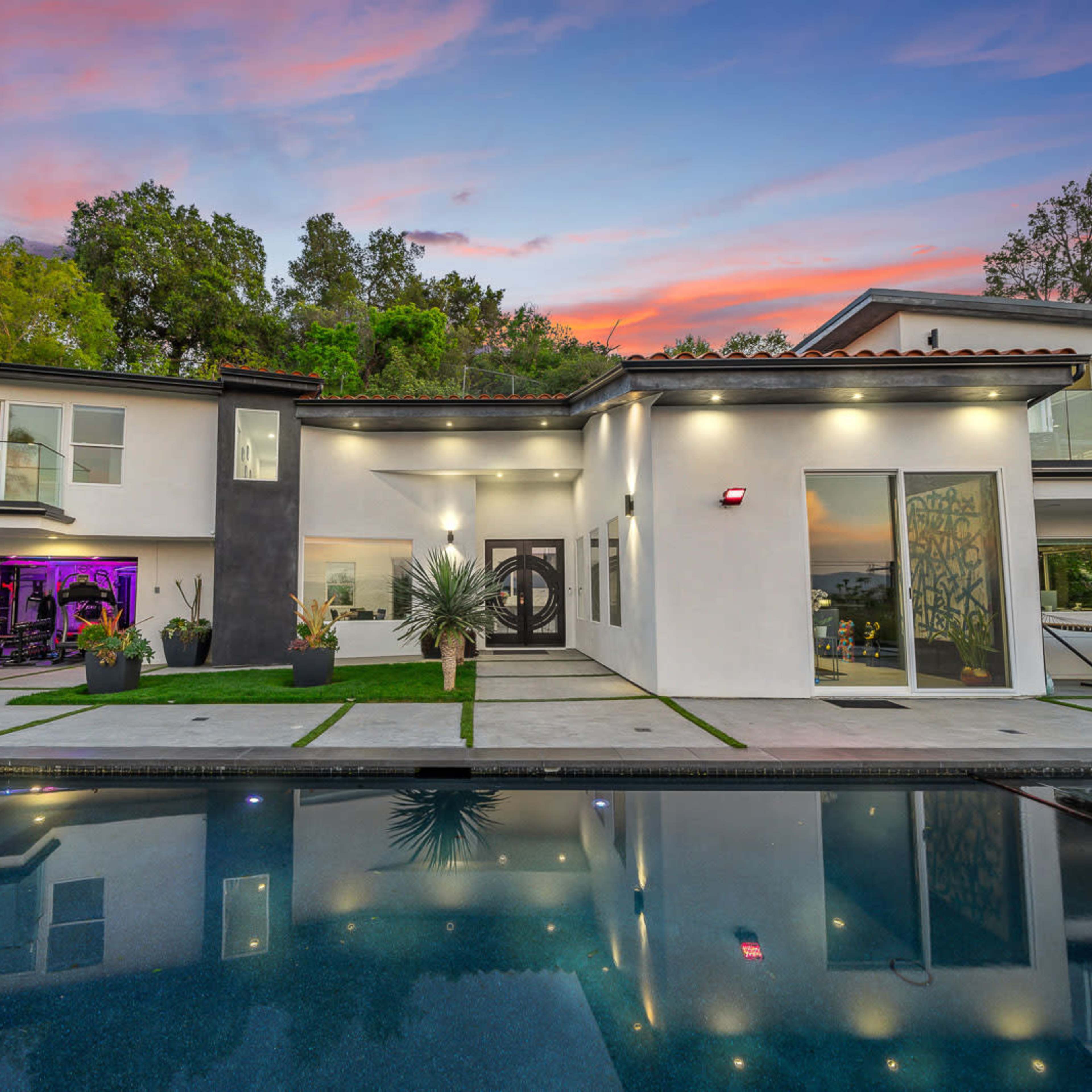 A modern two-story house with large glass windows and a pool, set against a colorful sunset sky.