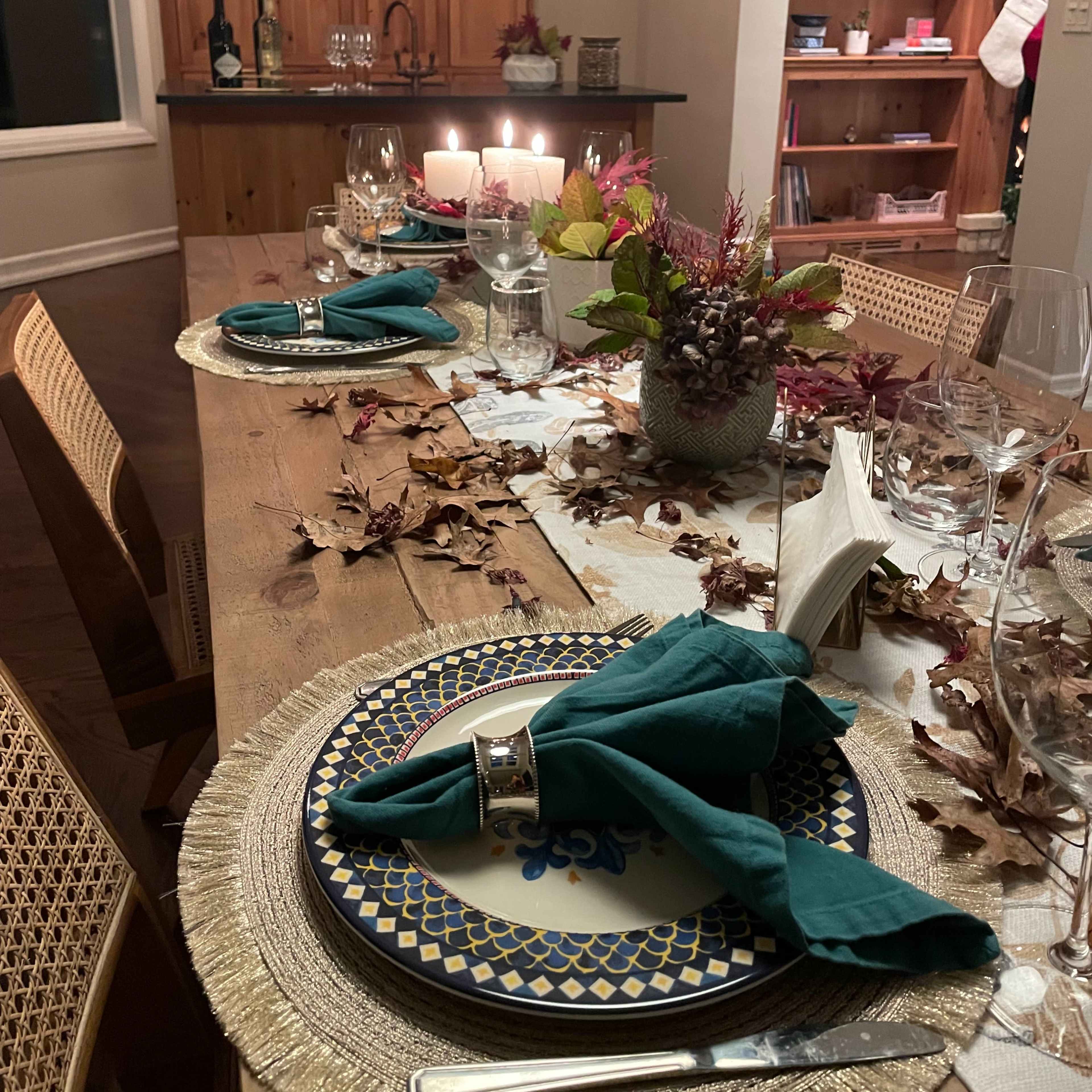 The dining table is set with plates and glassware, surrounded by fall leaves and candles, with a backdrop of wooden cabinets and a bookshelf.
