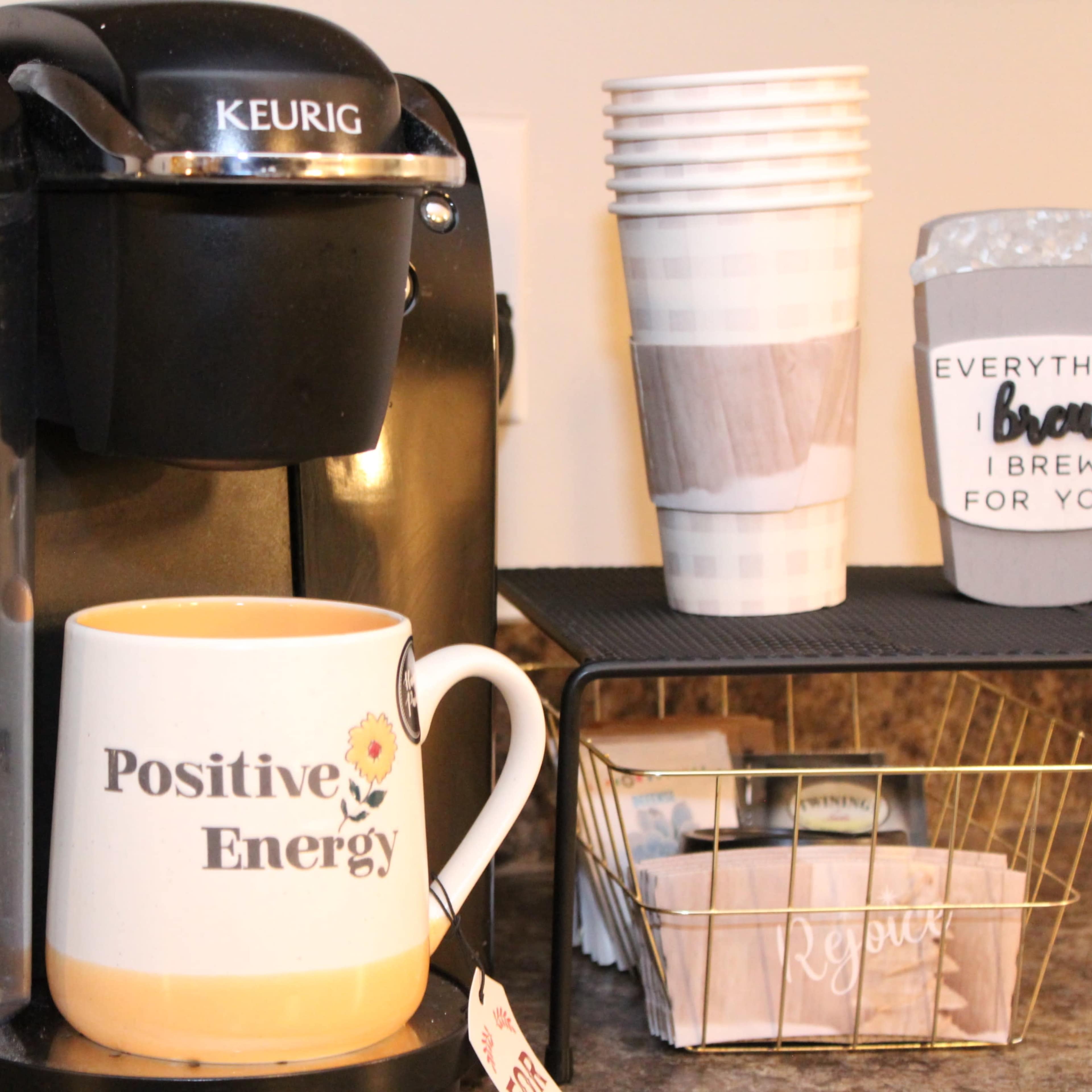A Keurig coffee maker is positioned next to several stacked disposable cups and a wire basket containing tea bags.