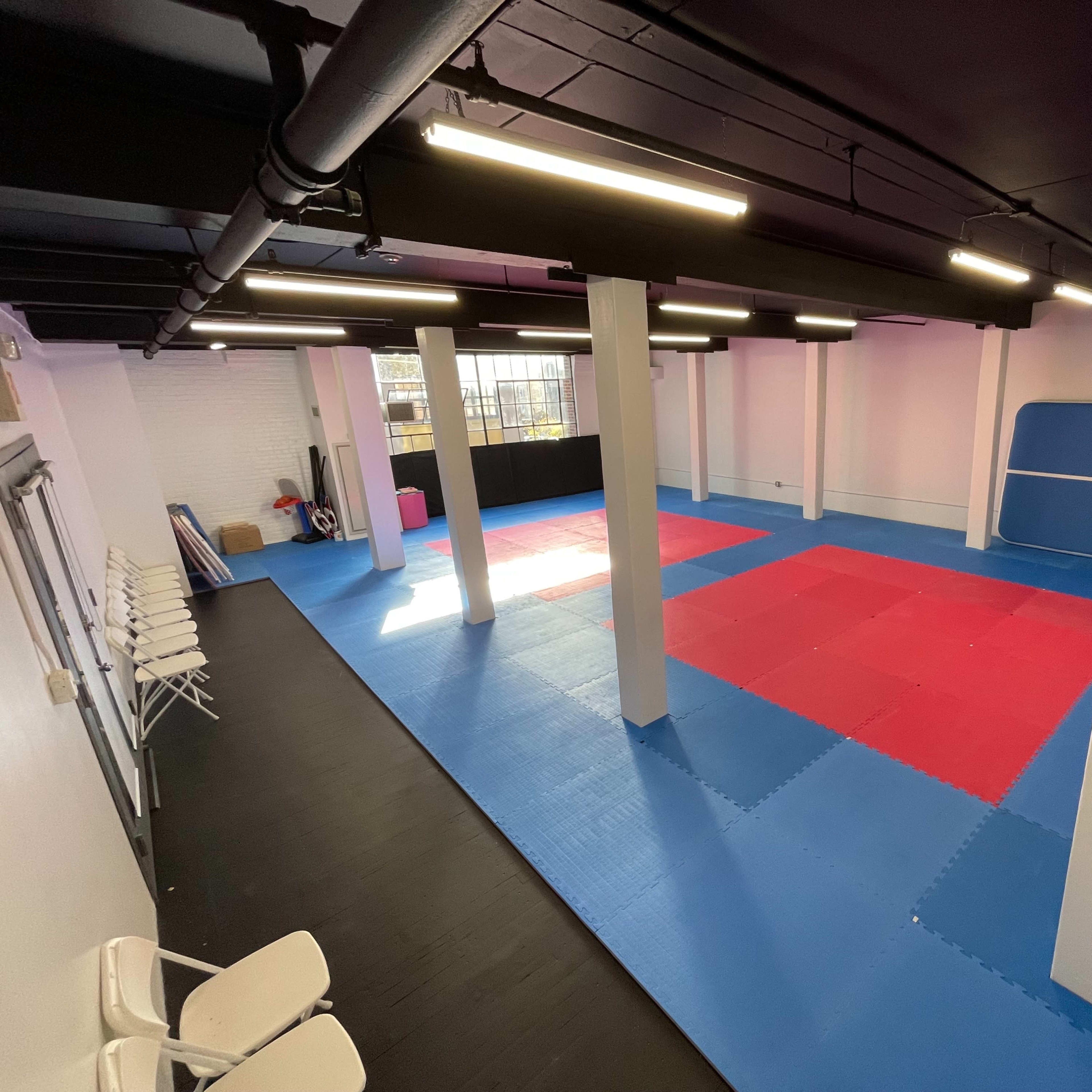 A spacious martial arts training room with red and blue mats, white columns, and overhead lighting.