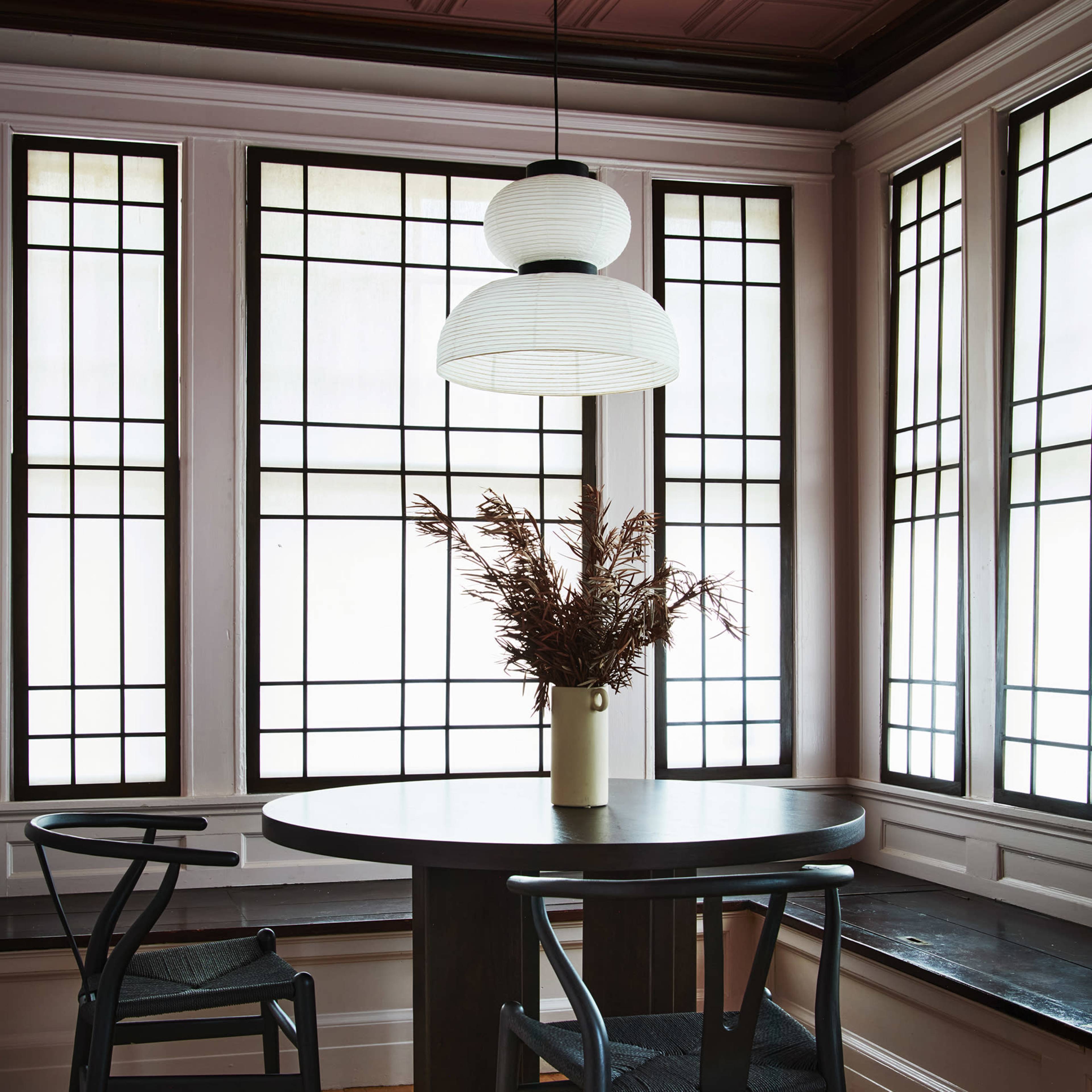 A round black dining table is positioned between two black chairs under a white pendant light, with a vase of dried pampas grass on the table, all set against a backdrop of large, grid-patterned windows.
