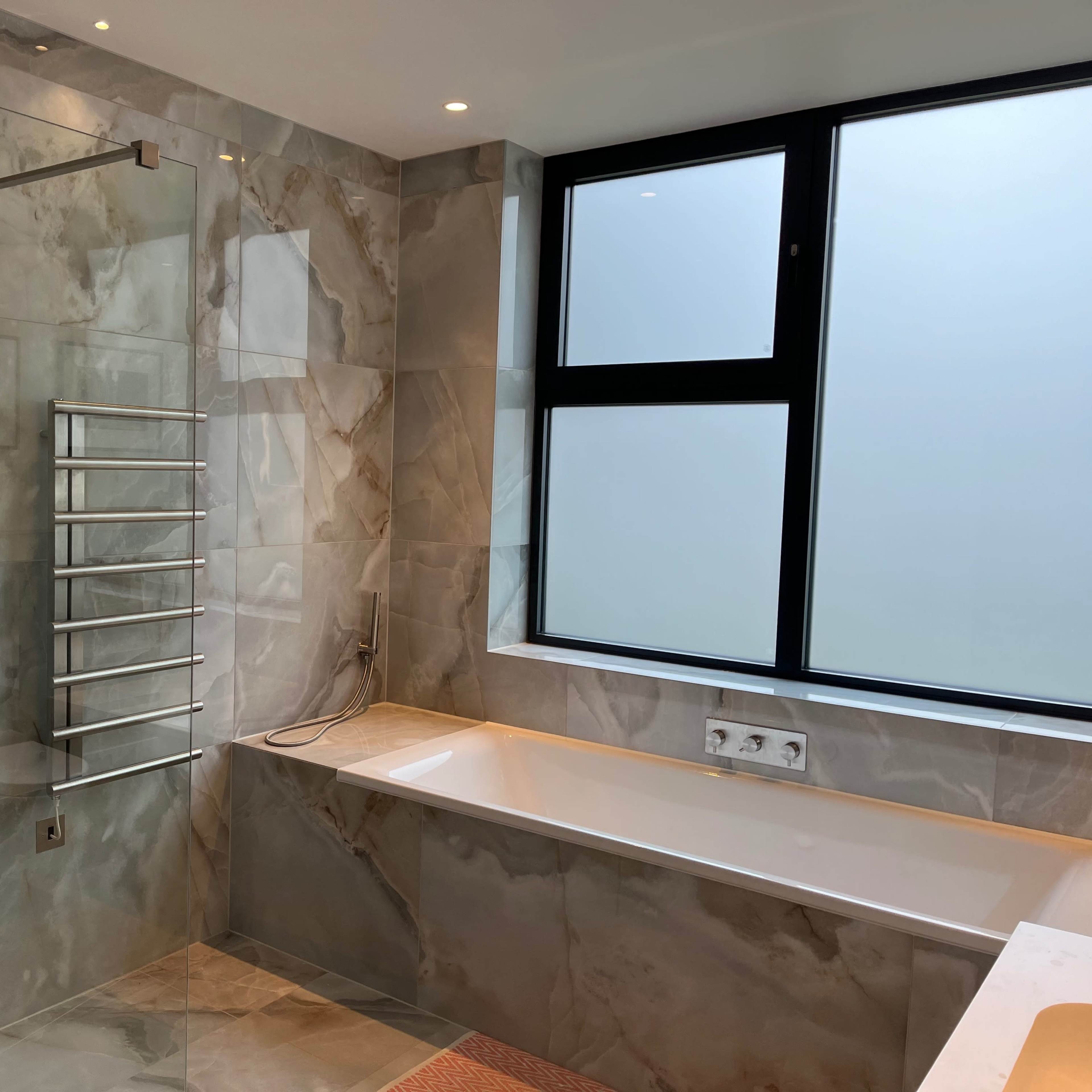 A modern bathroom featuring marble walls, a glass shower enclosure, a bathtub, and a large window.