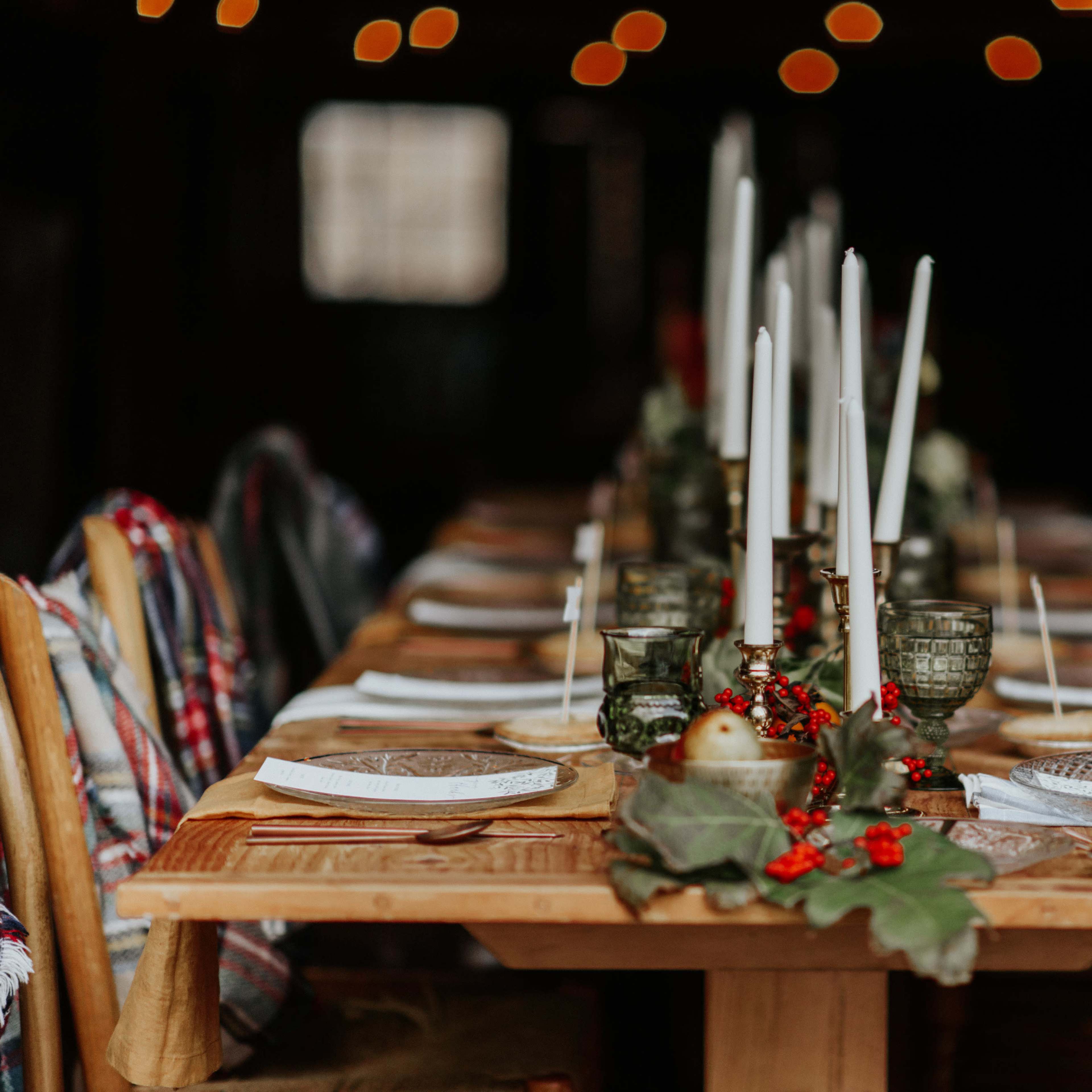A long wooden dining table is set with plates, candles, and greenery, surrounded by chairs draped with plaid blankets.