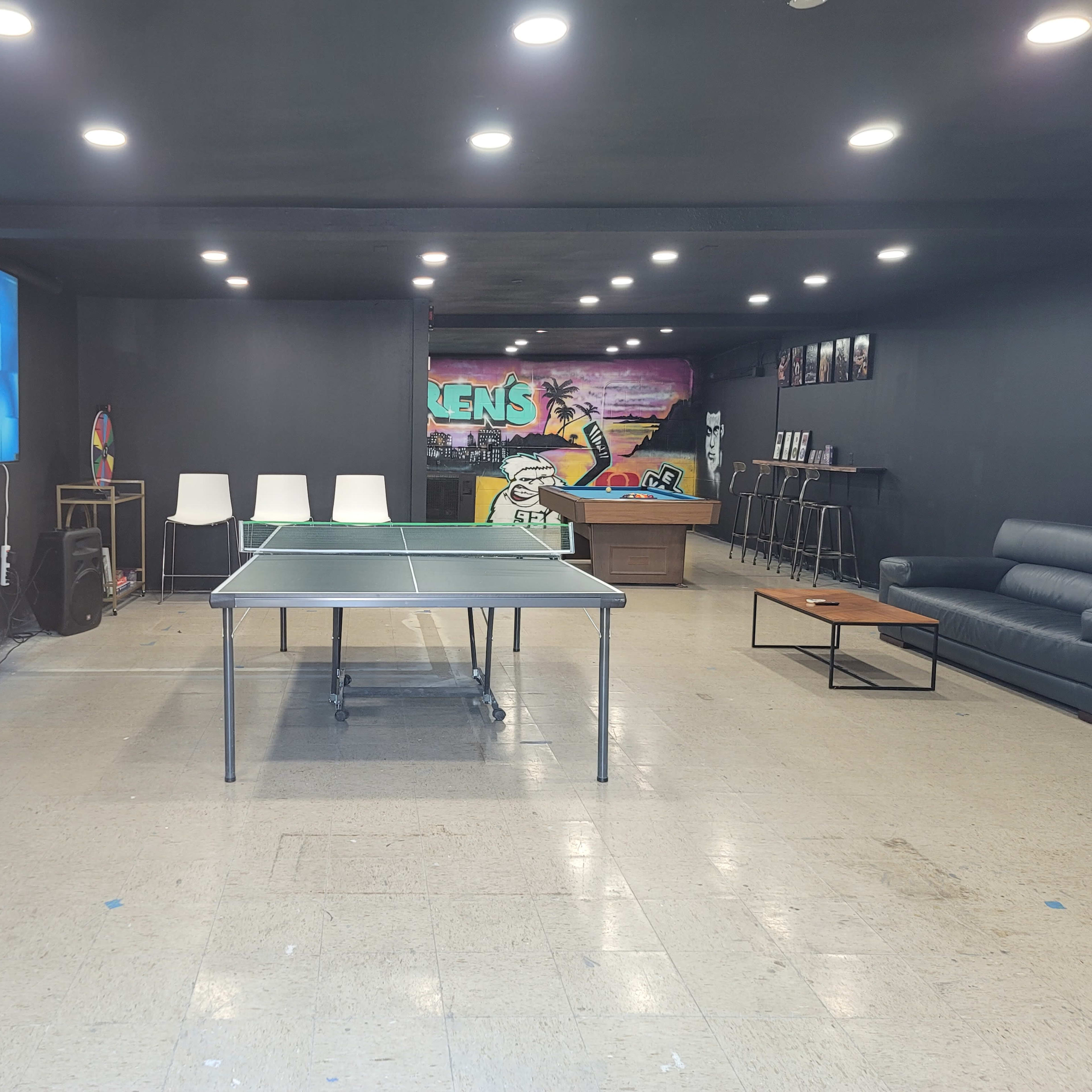 A room featuring a ping pong table, white chairs, a couch, a television screen, and a colorful mural on the wall.