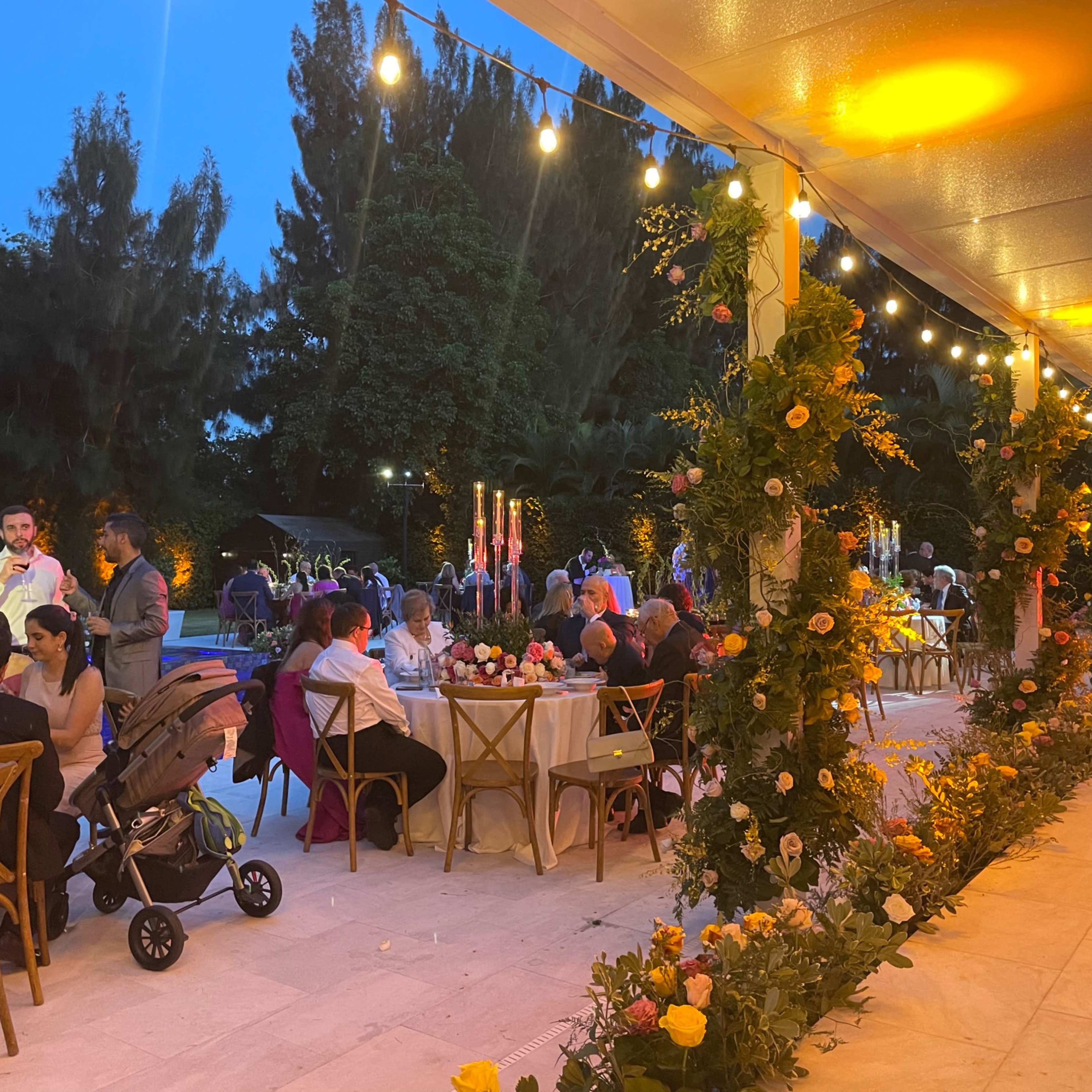 A well-decorated outdoor venue features tables with floral arrangements as guests dine and socialize under string lights during dusk.