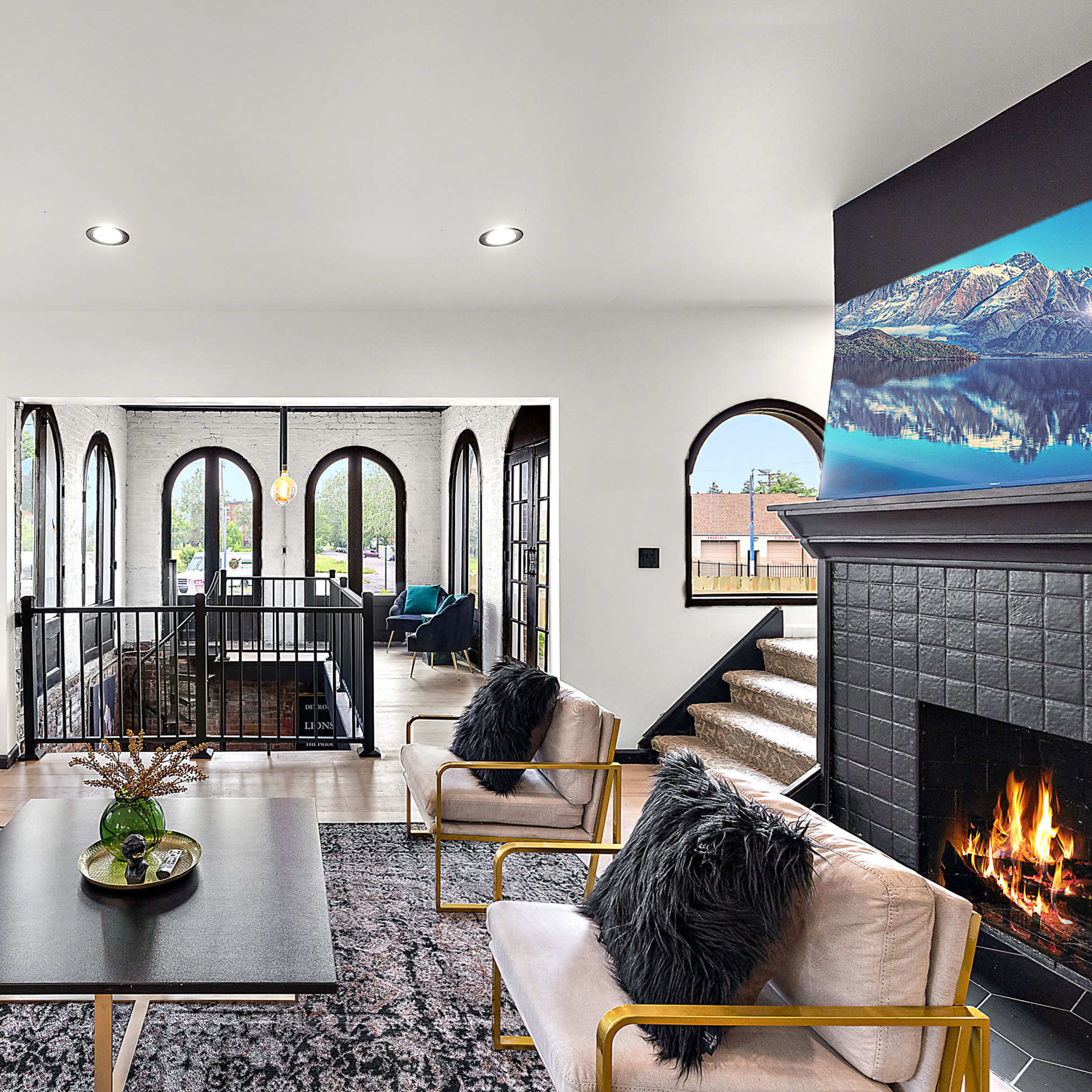 A modern living room with a teal sofa, plush armchairs, a coffee table, and a fireplace, complemented by arched windows and a view of a staircase leading to another room.