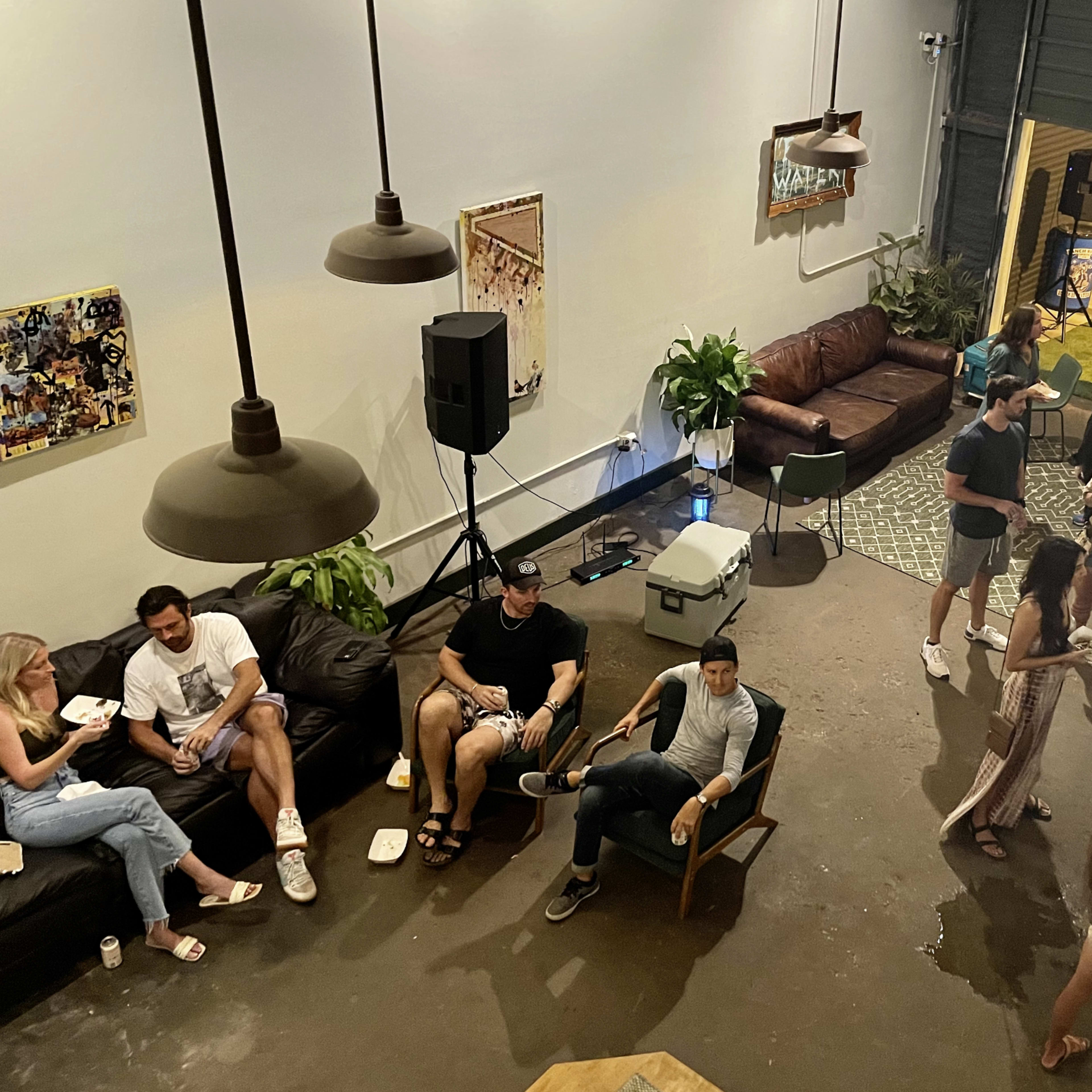 A group of people socialize in a spacious indoor venue with a mix of seating arrangements and a visible outdoor area.