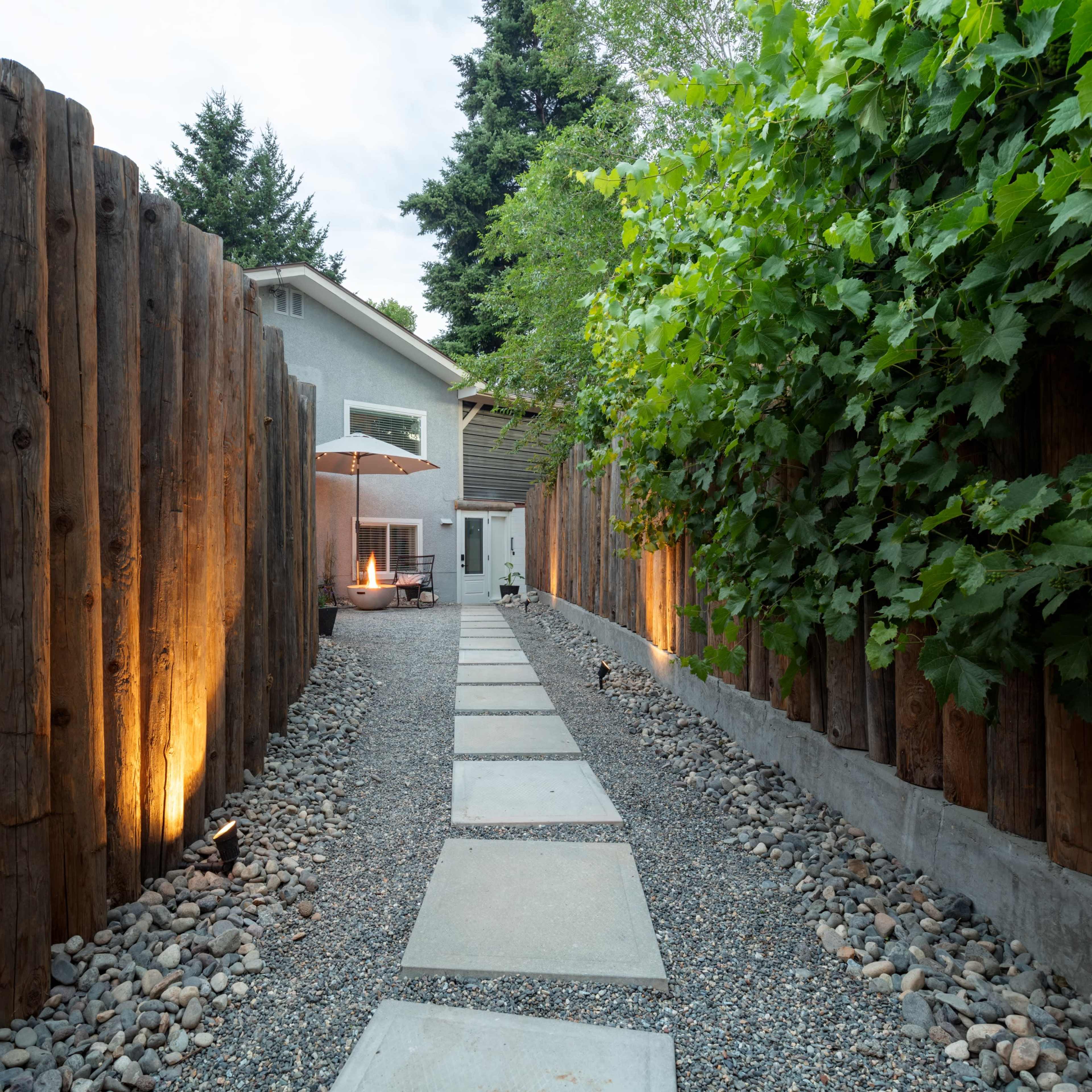 A gravel pathway lined with stepping stones leads to a modern house, enclosed by tall wooden fencing and greenery.