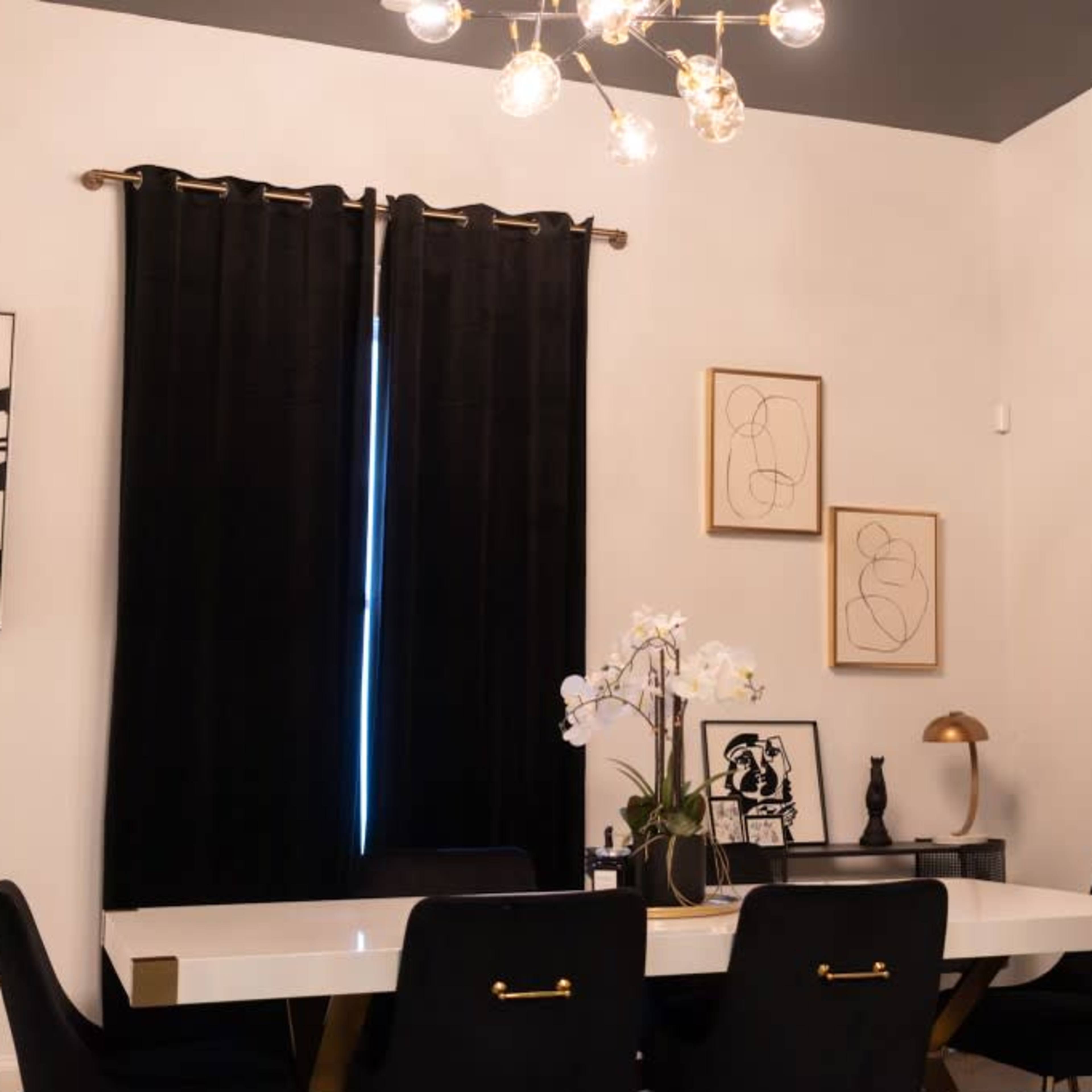 A modern dining area features a white table surrounded by black chairs, with black curtains and framed artwork on the walls.
