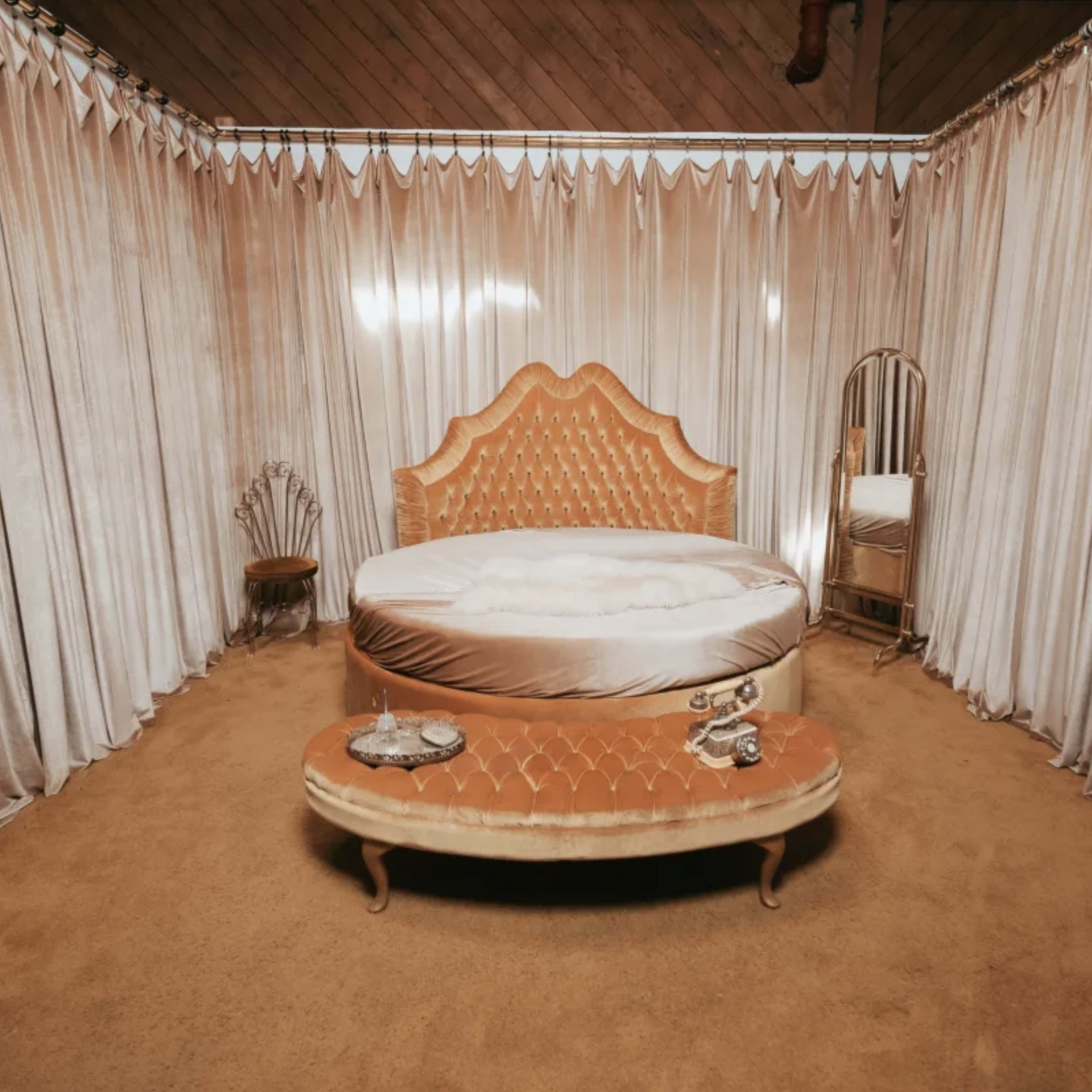 A round, tufted bed surrounded by floor-length fabric curtains in a softly lit room with beige carpeting.
