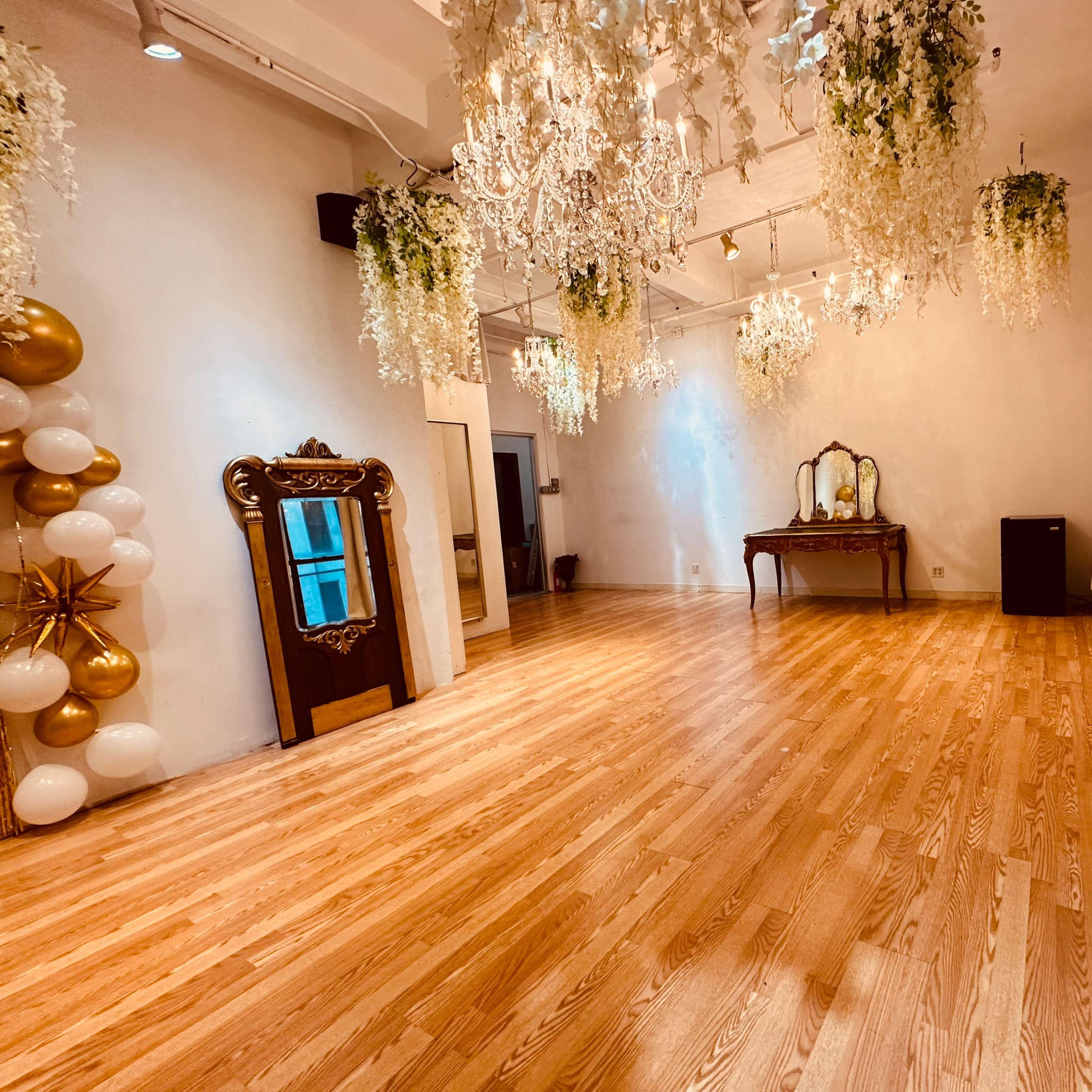 A spacious room with wooden flooring, adorned with chandeliers and hanging greenery, featuring a large mirror and a vintage-style table against one wall.