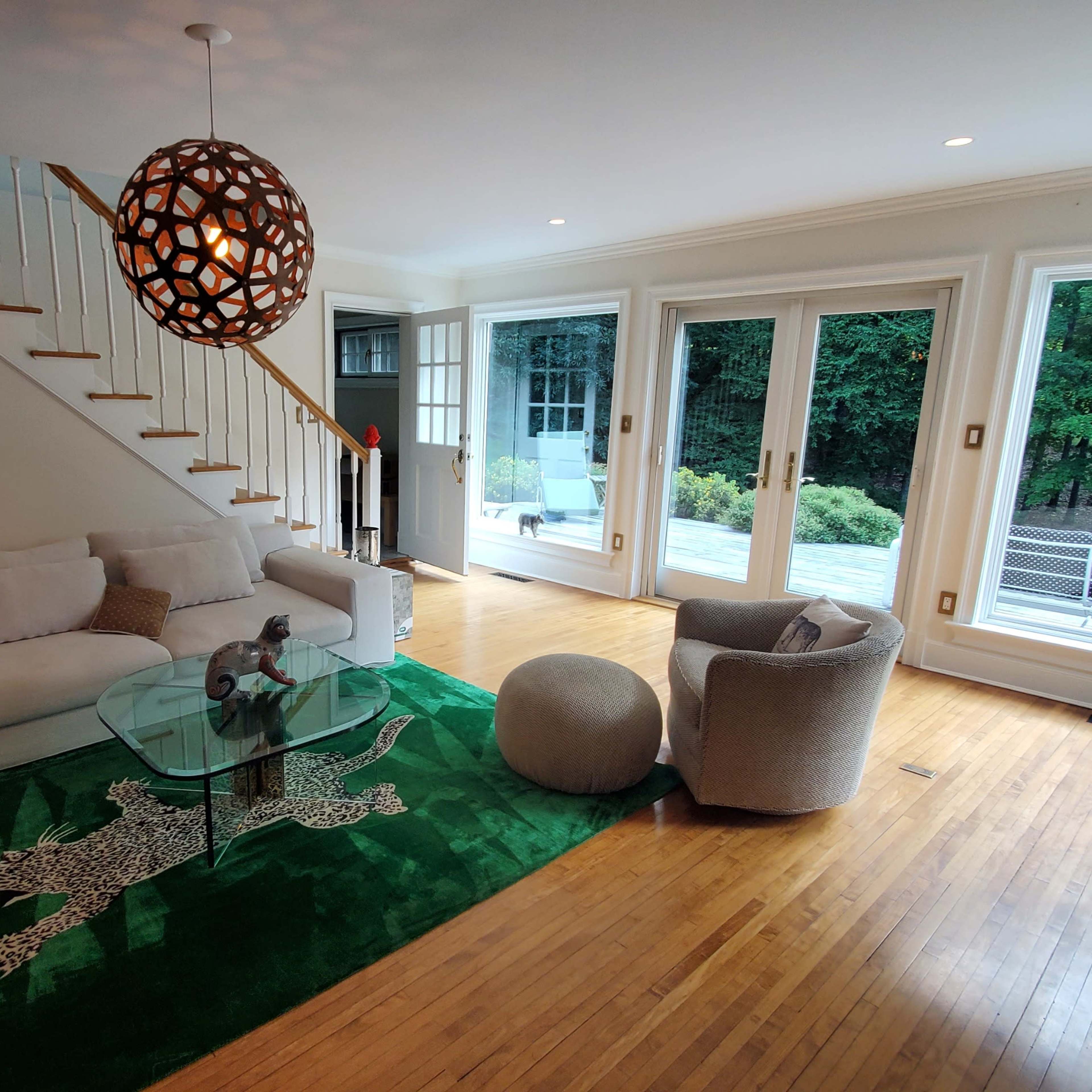 A living room features a white sofa, a circular glass coffee table, a green rug with a leopard print design, and large windows overlooking a patio.