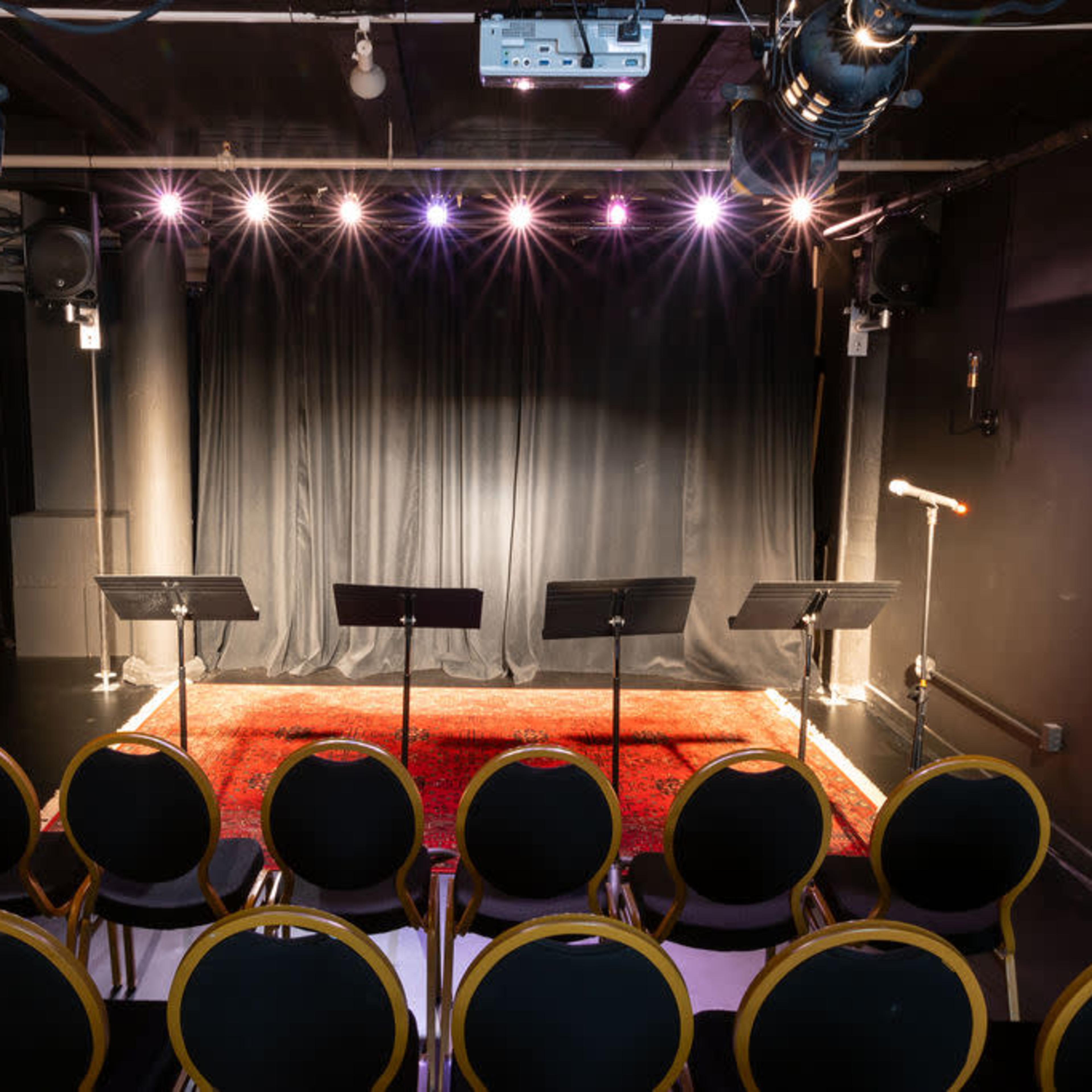 A small theater stage with a black curtain, spotlighted by overhead lights, and a red rug, facing a row of empty circular chairs.