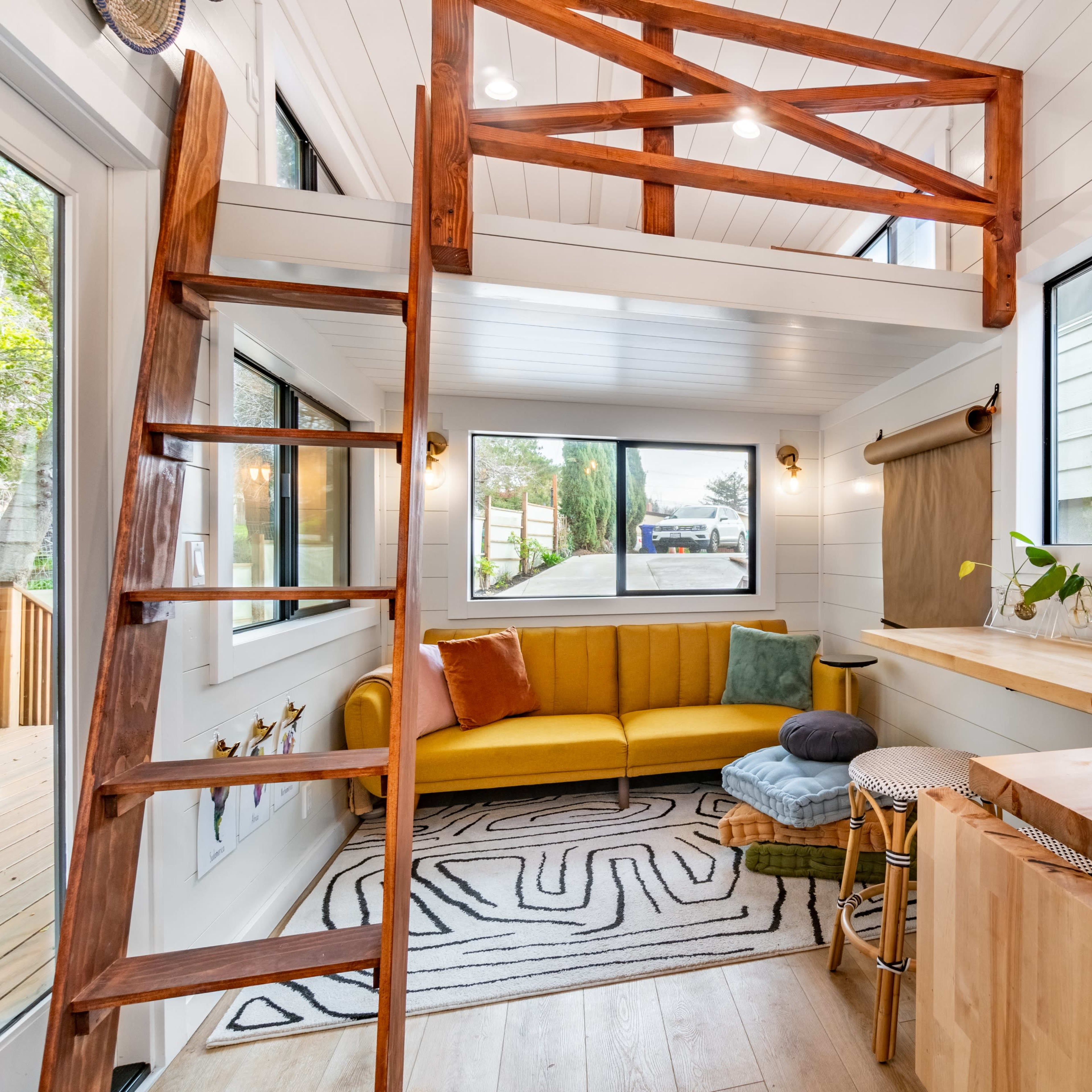 A bright, modern interior of a small living space featuring a yellow sofa, a wooden ladder leading to a loft area, and a small kitchen bar with stools.