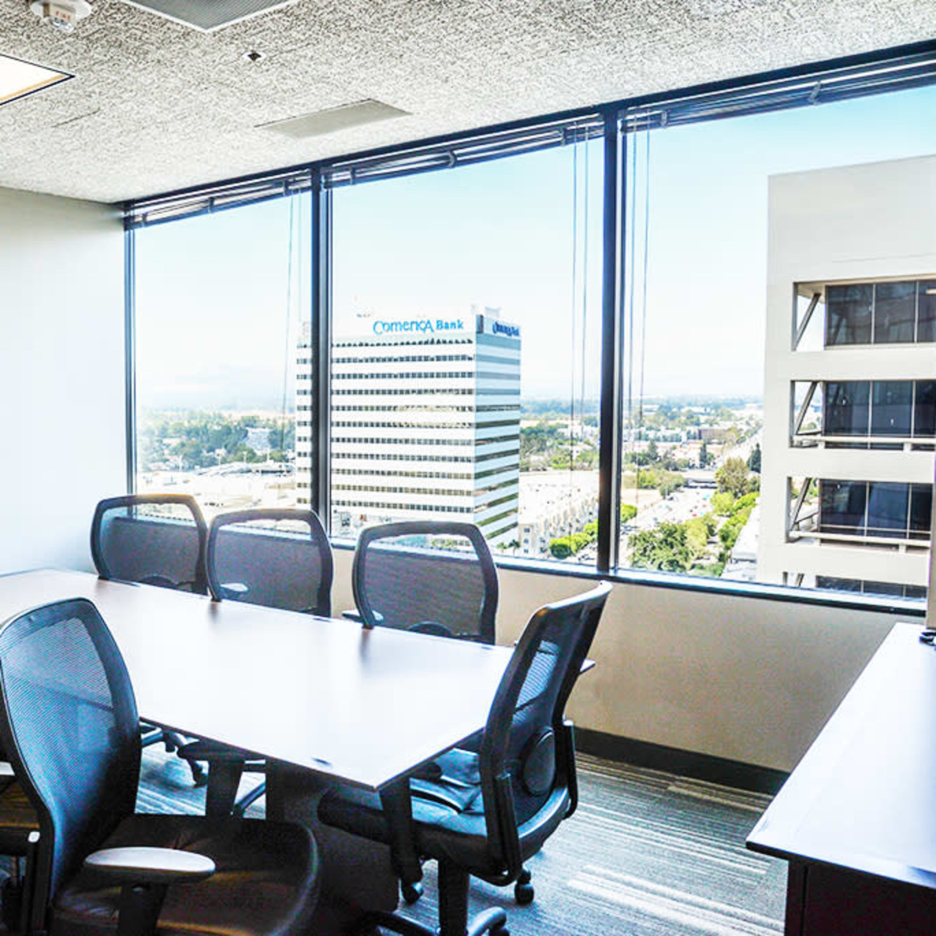 A conference room with a long table and several mesh chairs features large windows displaying an urban view of buildings outside.