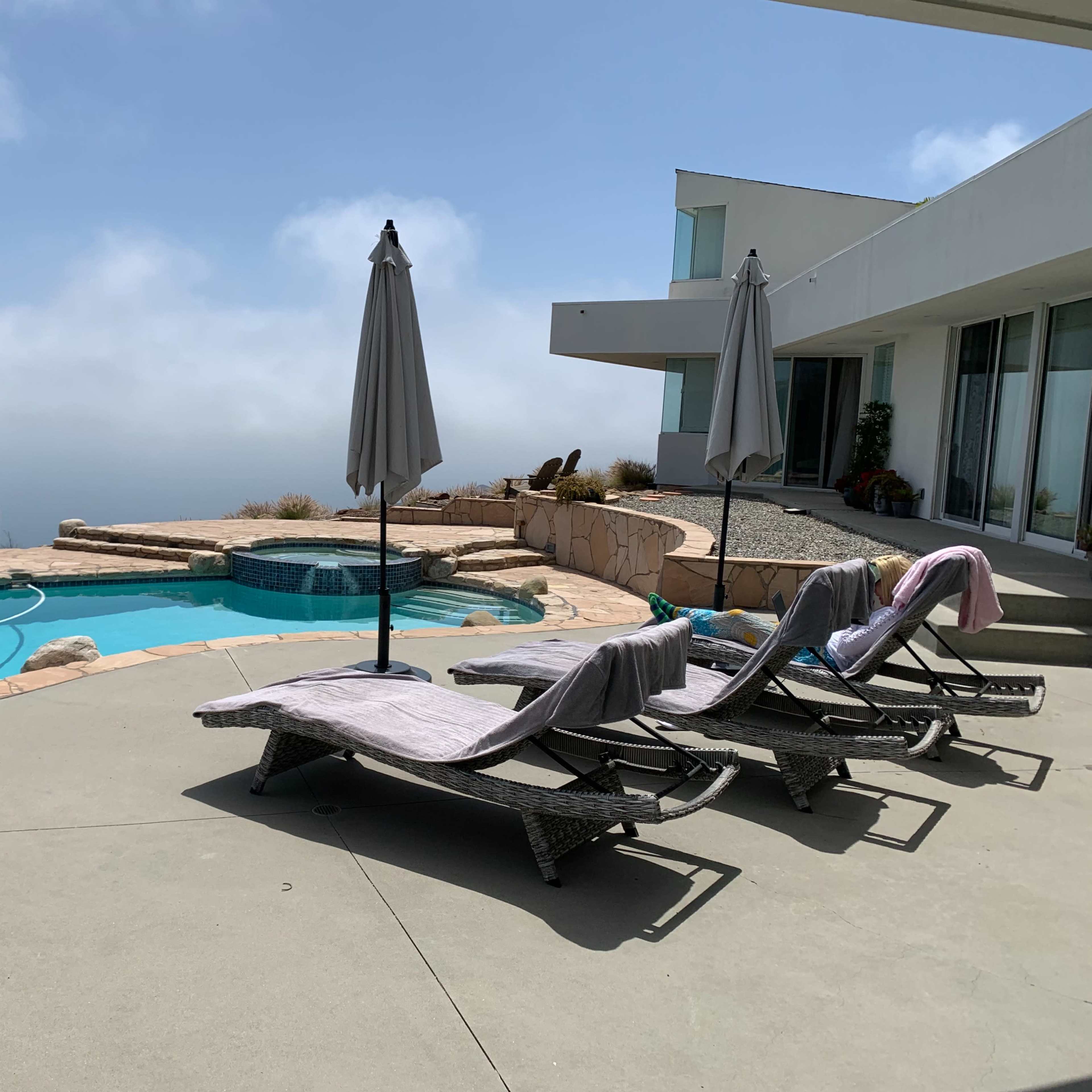A swimming pool area with two lounge chairs and umbrellas, overlooking a distant foggy landscape.