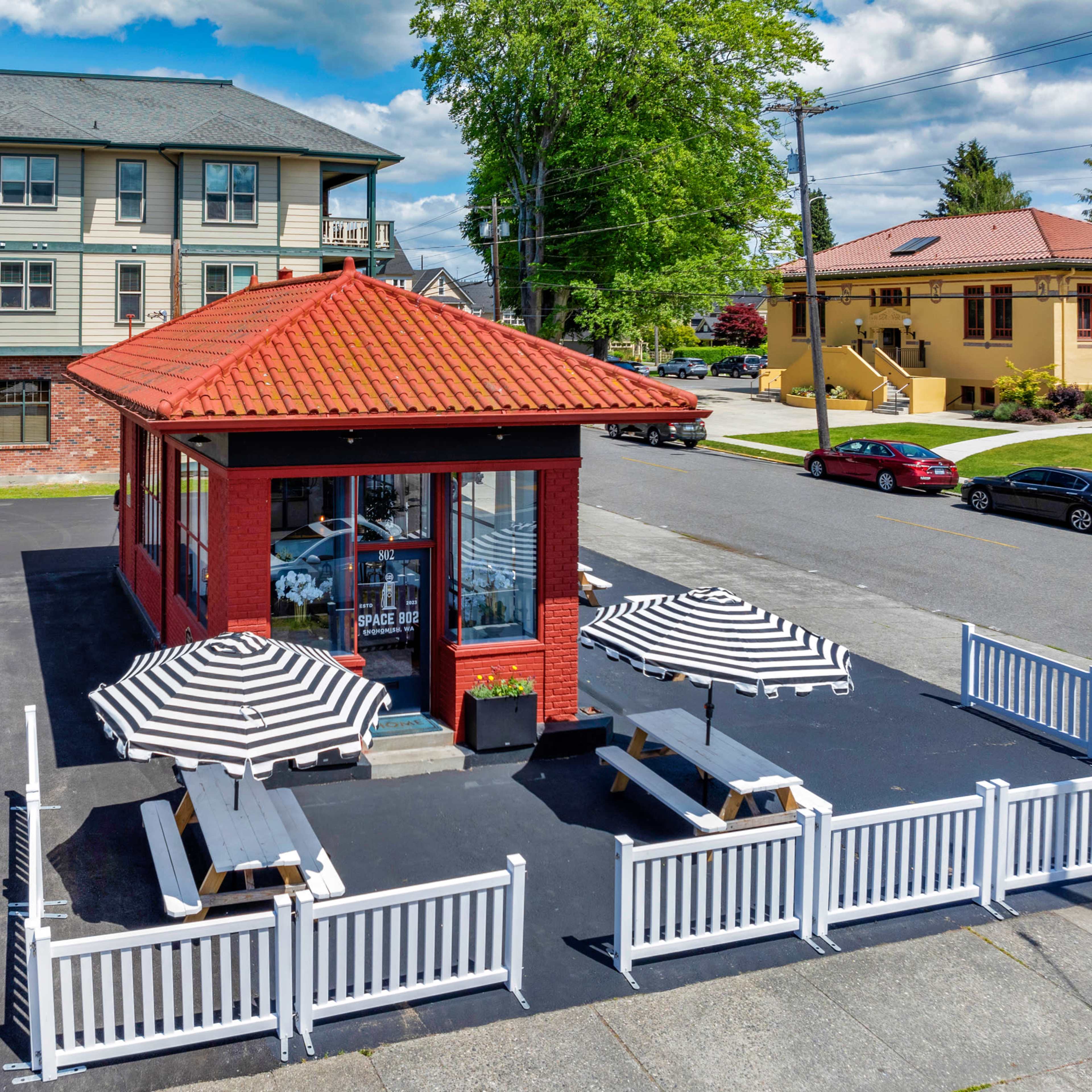 A small red brick building with a tiled roof, surrounded by white fencing and featuring striped umbrellas over outdoor seating areas.