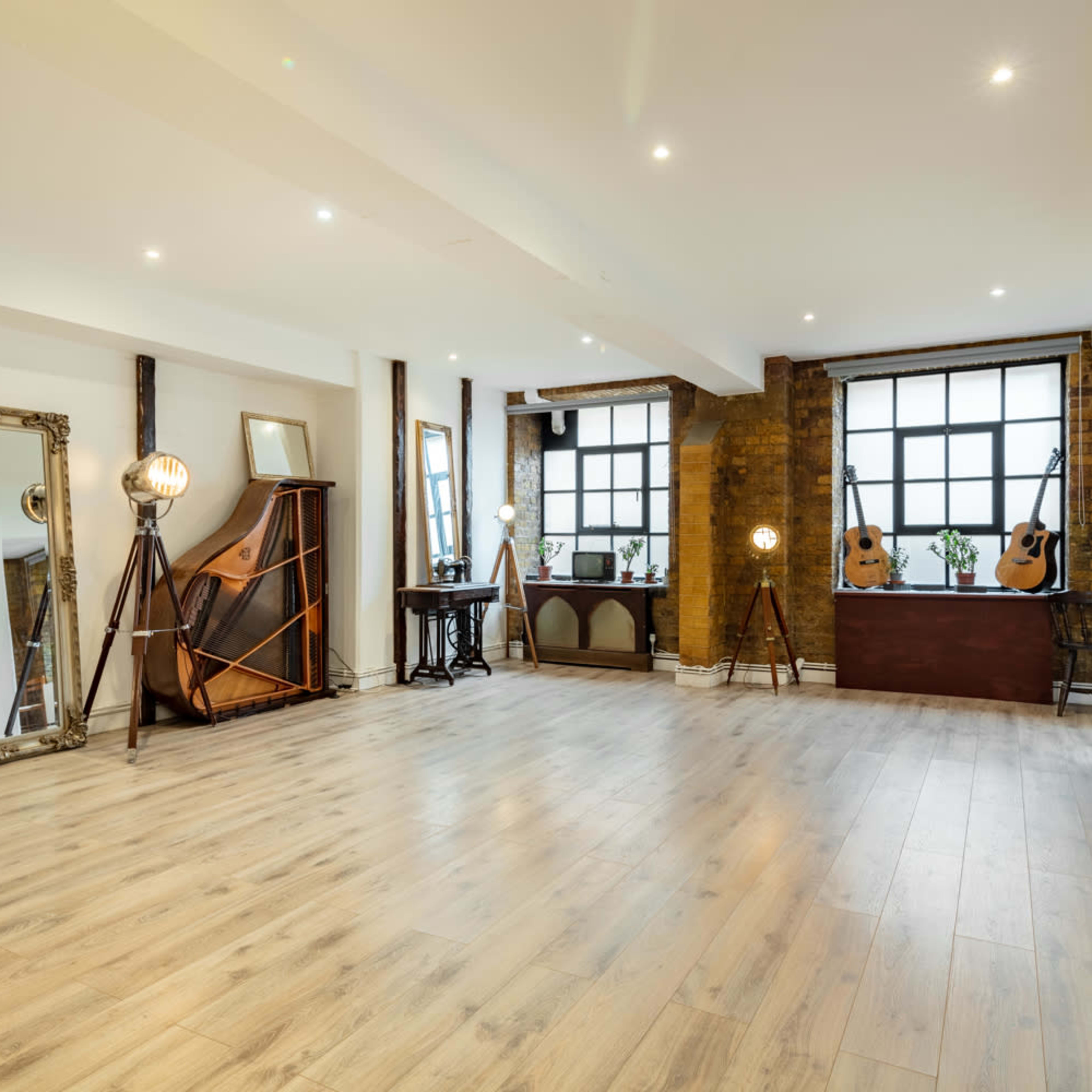 A spacious room features wooden flooring, large windows, and a mix of decorative furniture, including mirrors and guitars.