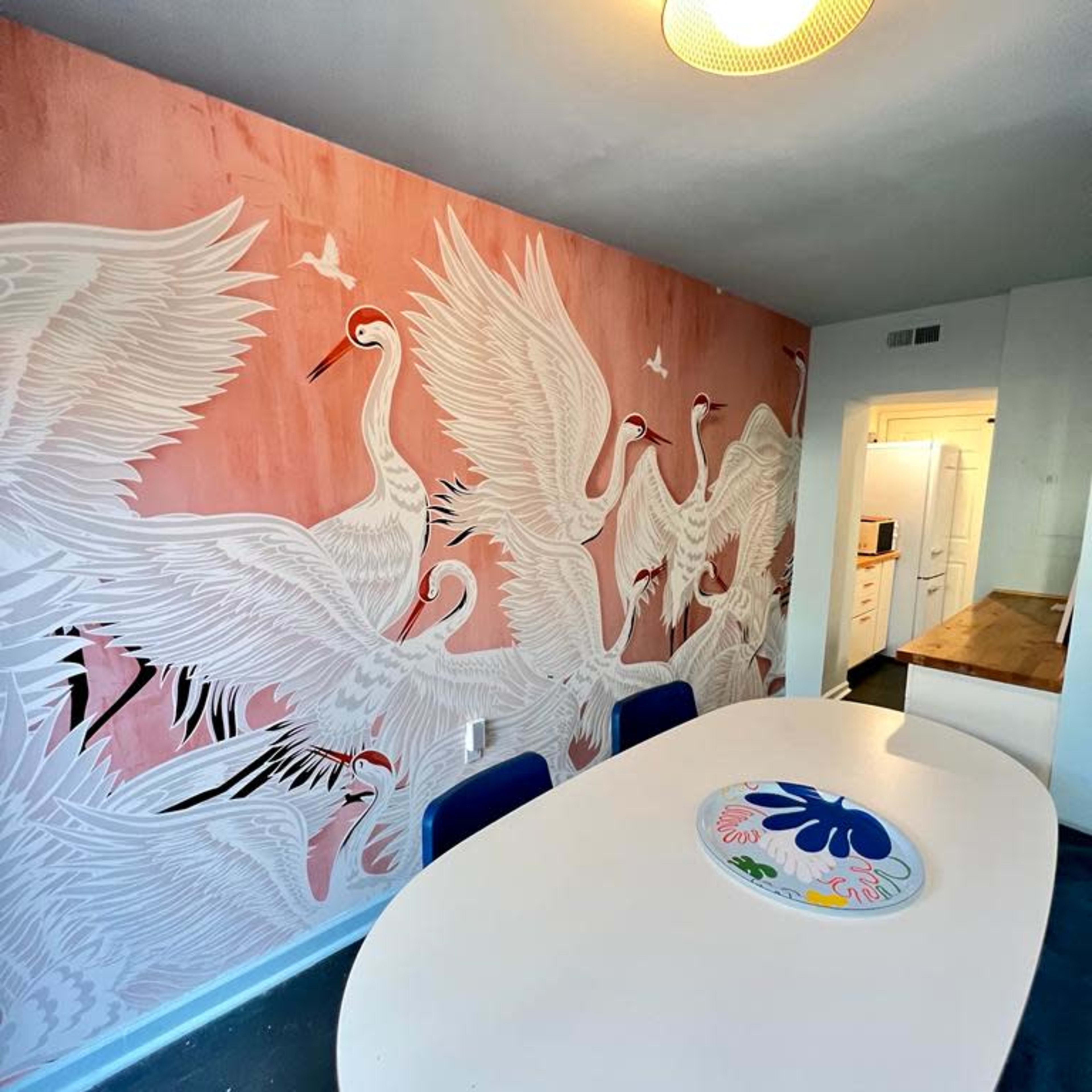 A dining area with a large oval table surrounded by chairs, set against a pink wall adorned with a mural of white cranes in flight.