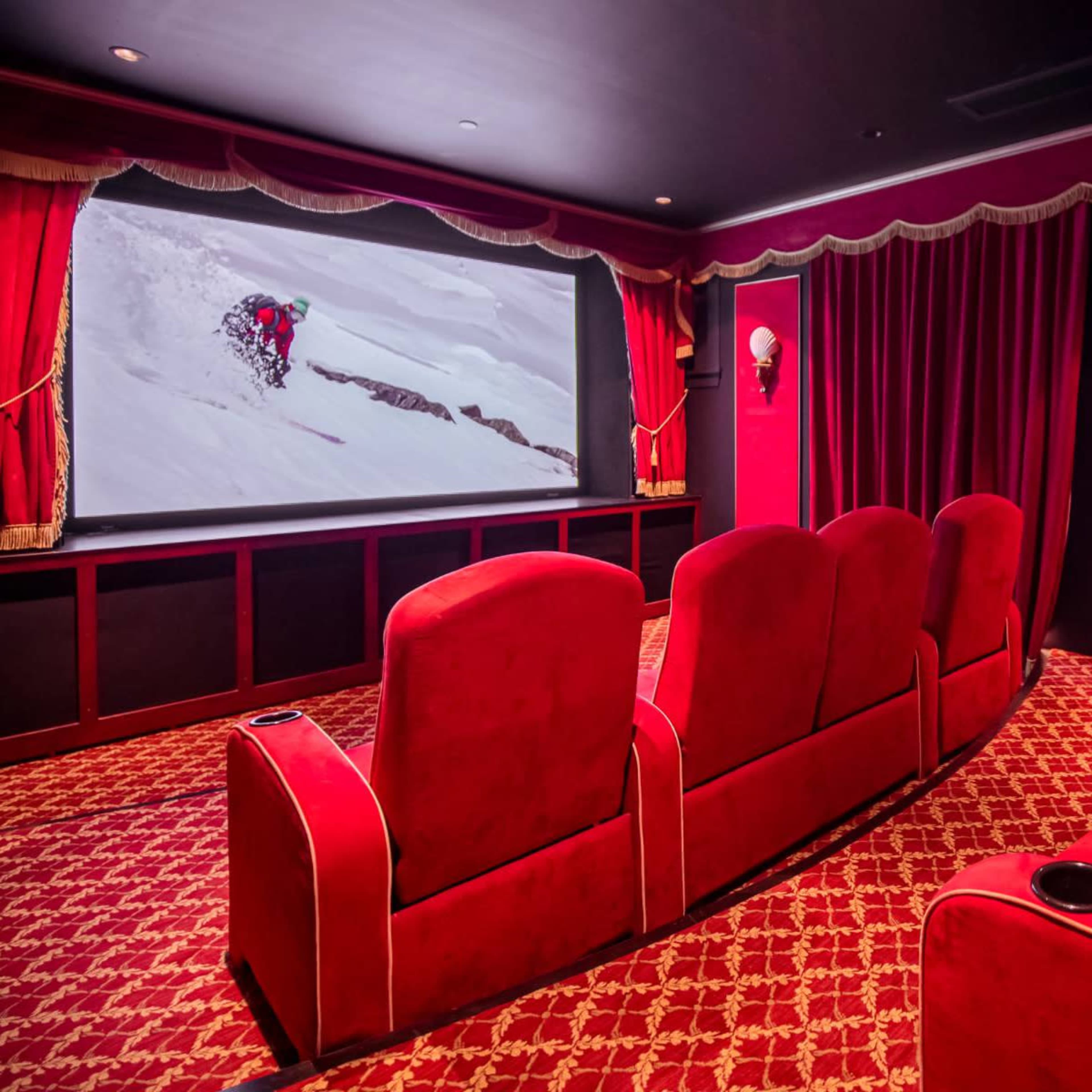 A private home theater with red velvet seating and draped curtains, featuring a large screen displaying a skiing scene.