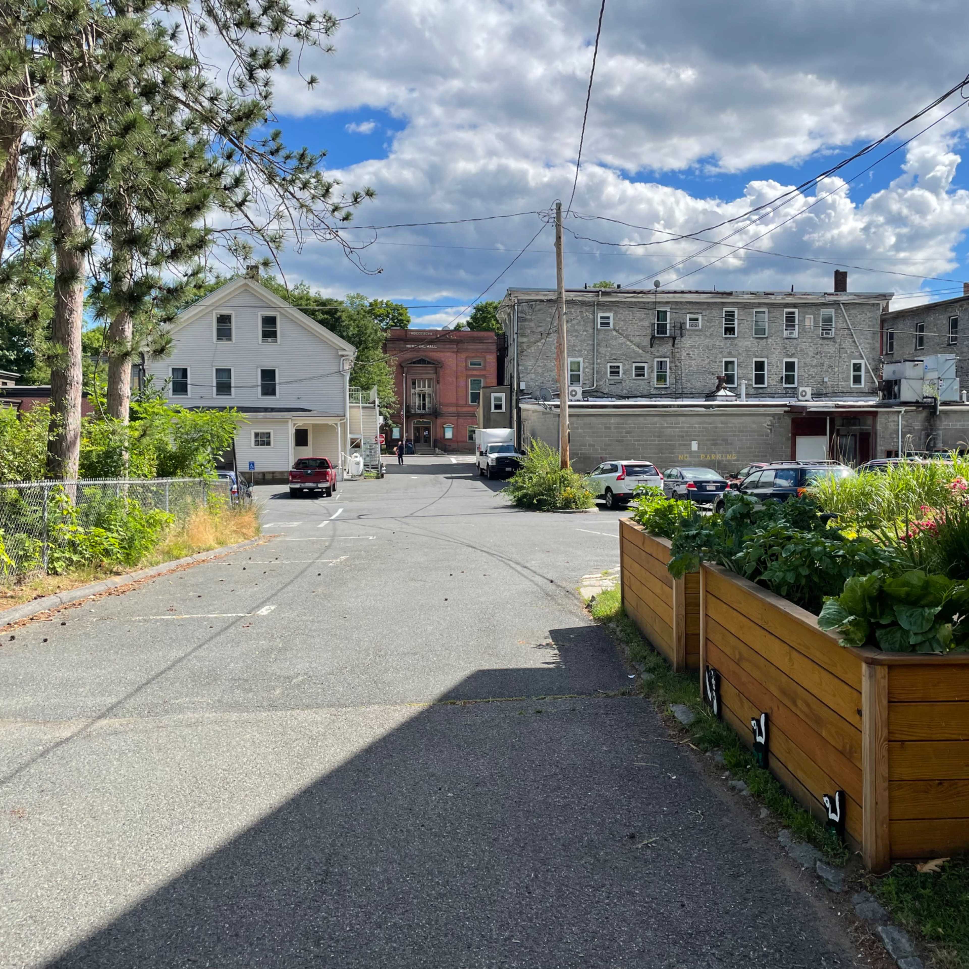 A narrow alleyway is lined with greenery and leads to a gravel road, with several parked cars and buildings in the background under a partly cloudy sky.