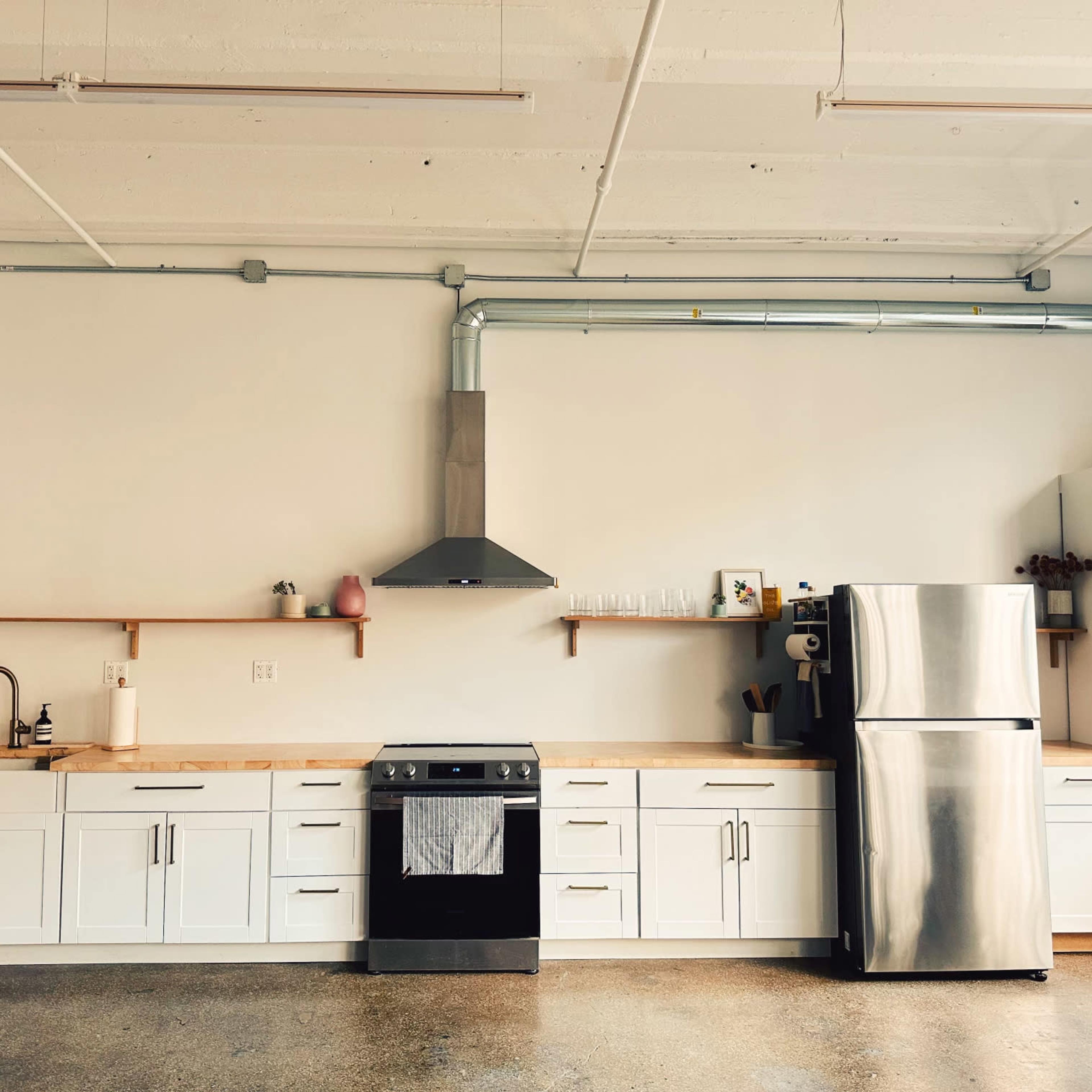 A modern kitchen features white cabinetry, a stainless steel refrigerator, and a stove with an overhead exhaust hood on a concrete floor.